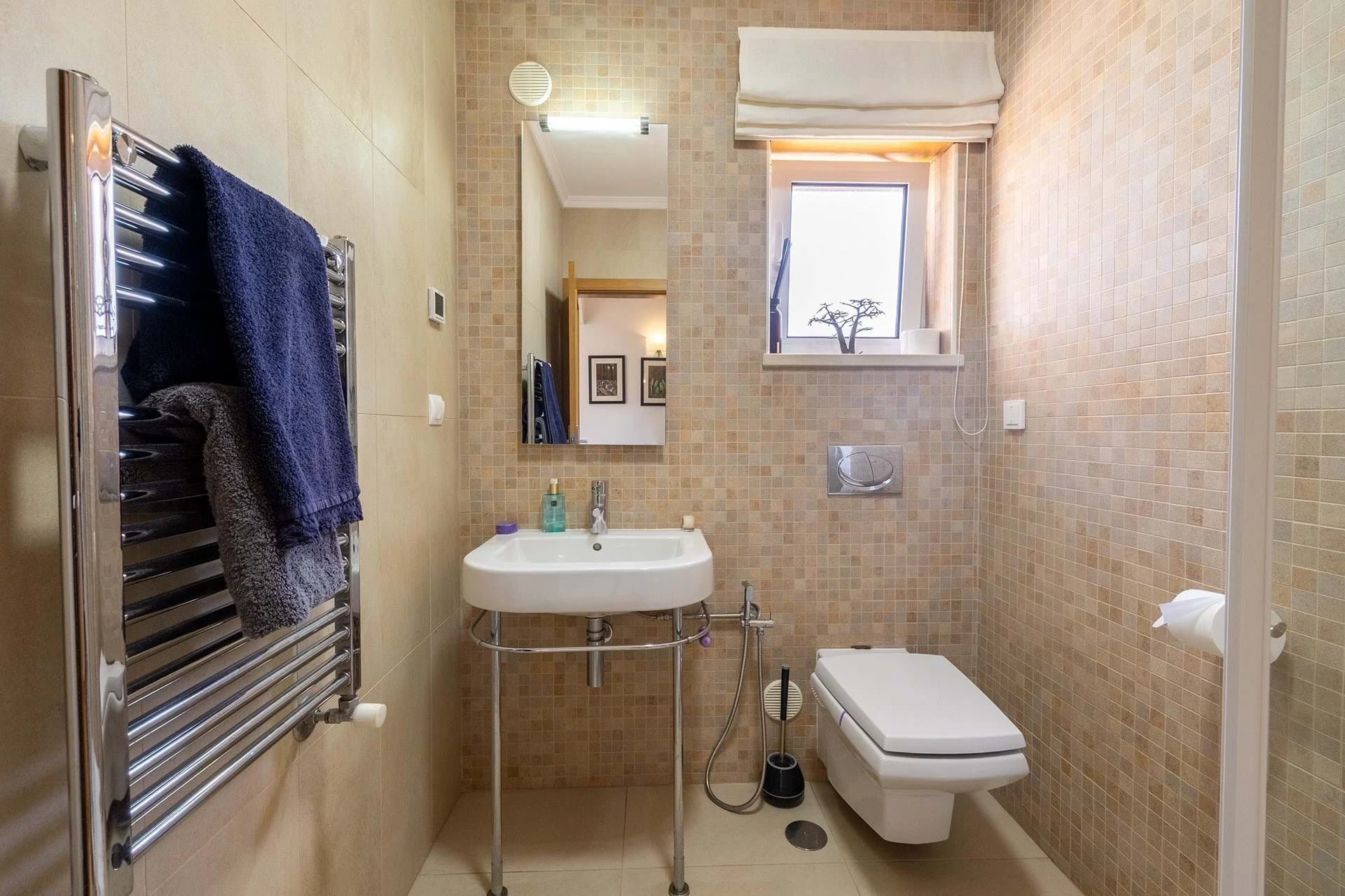 T5 Villa with pool in Ferrel 5mn from beach and Lagos centre – SunnySteve 25 Small modern bathroom with a wall-hung toilet, white sink, beige mosaic walls, and chrome towel warmer with a navy towel nearby.