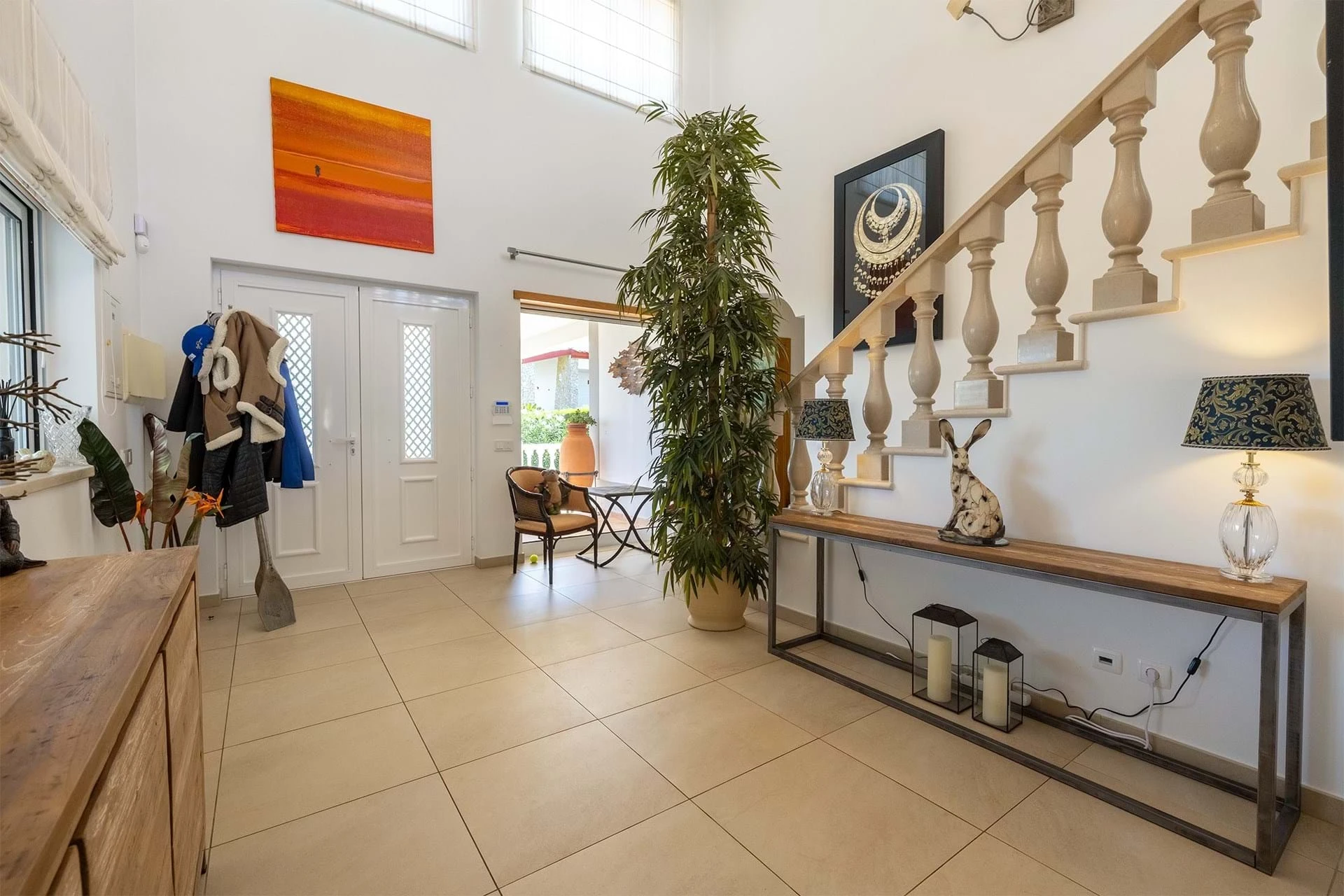 T5 Villa with pool in Ferrel 5mn from beach and Lagos centre – SunnySteve 27 Bright entry hall with a tall plant near a wooden staircase, a coat rack by the door, and a console table with lamps and decor on the wall side.