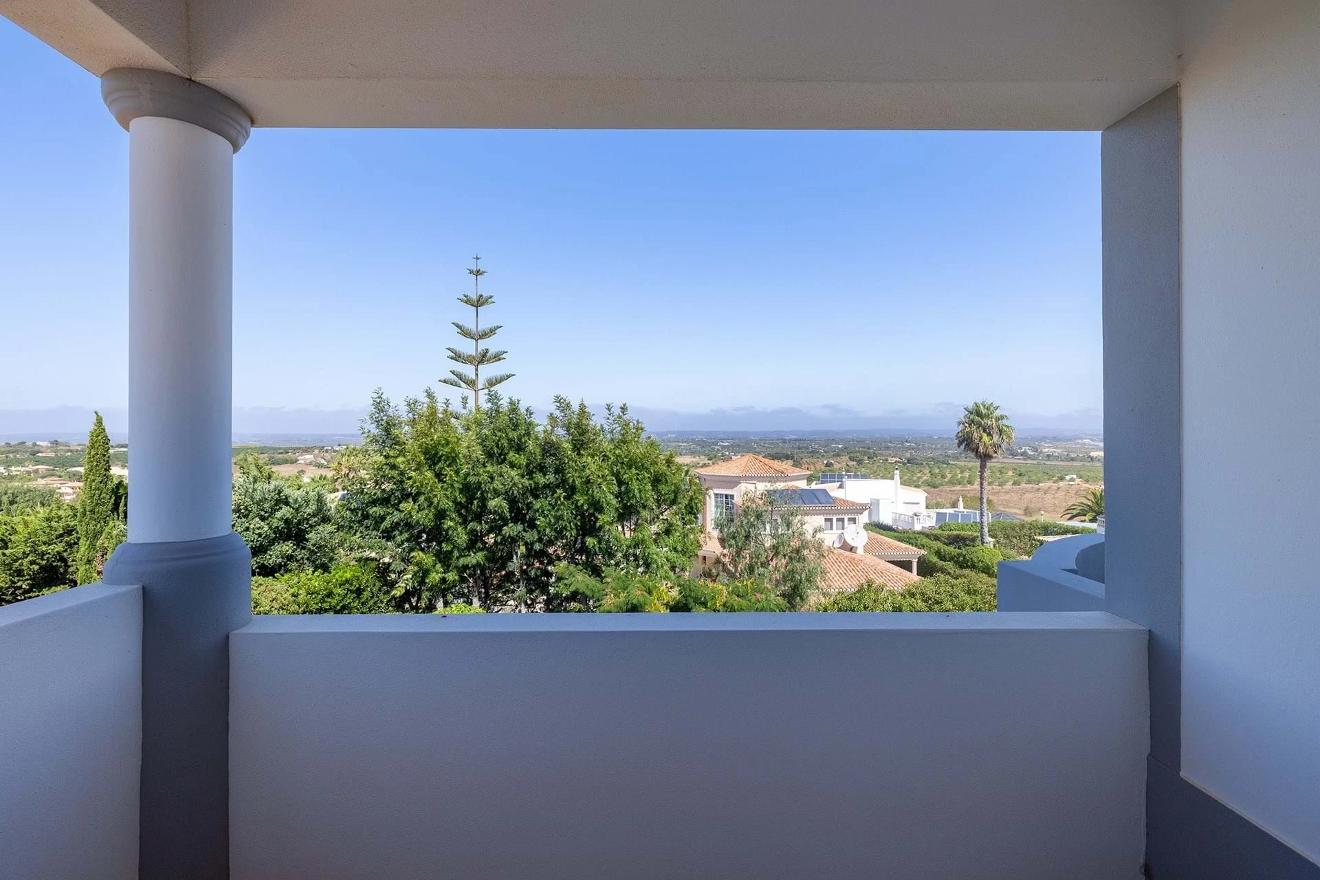 T5 Villa with pool in Ferrel 5mn from beach and Lagos centre – SunnySteve 30 View from a white balcony frame overlooking a suburban landscape with trees and distant hills under a clear blue sky.