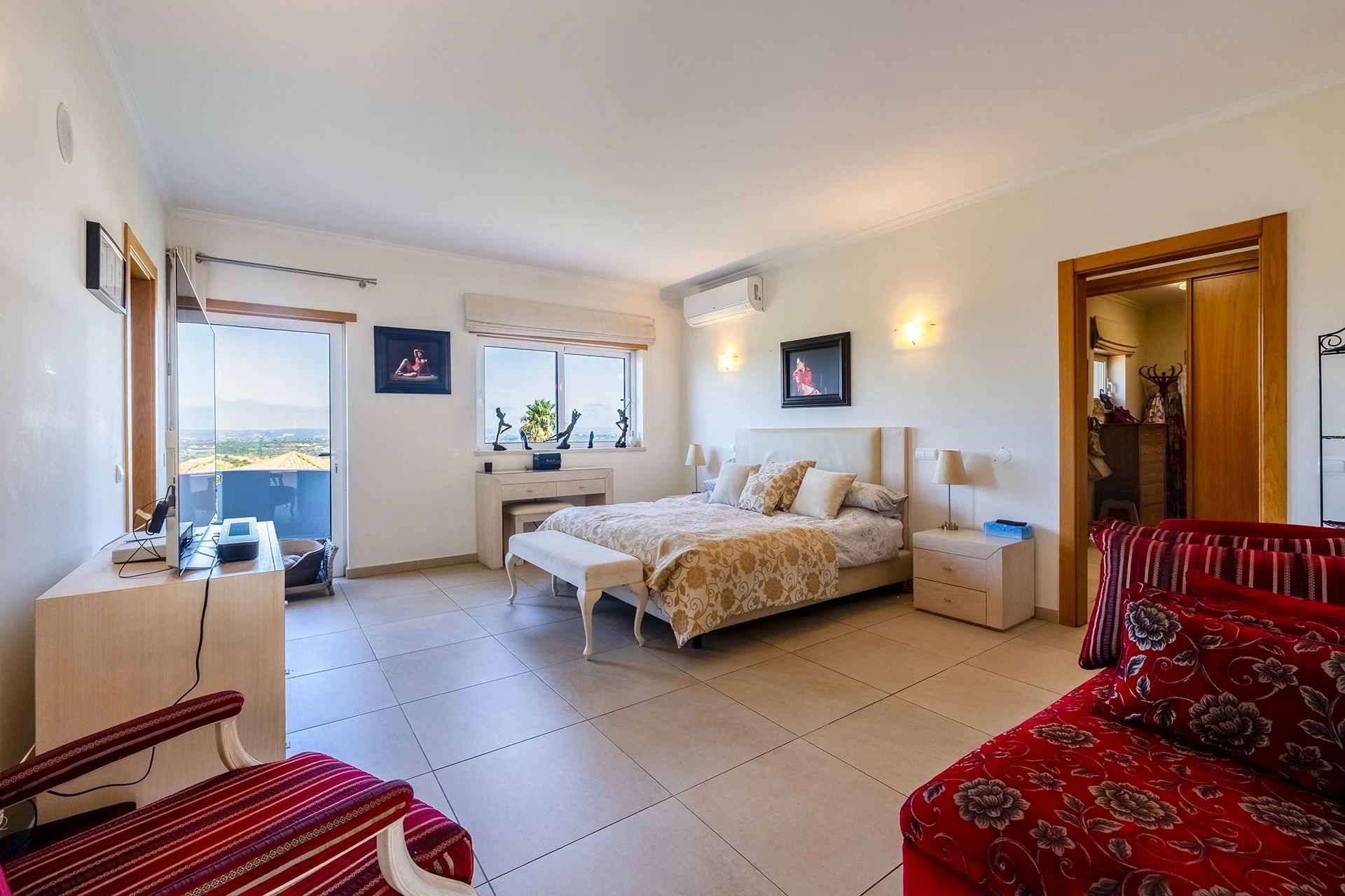 T5 Villa with pool in Ferrel 5mn from beach and Lagos centre – SunnySteve 34 Bright hotel bedroom with a large bed, beige bedding, and two matching nightstands; red floral chairs in the foreground; balcony and city view through glass doors to the left.