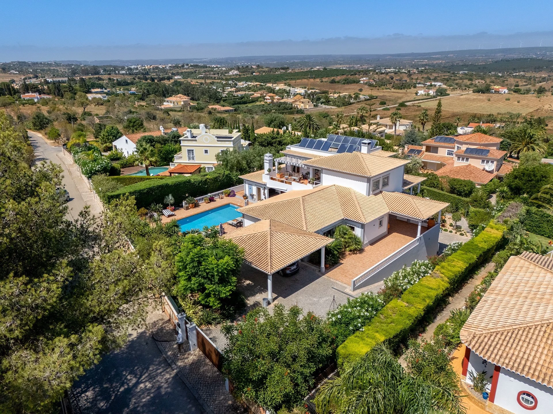 T5 Villa with pool in Ferrel 5mn from beach and Lagos centre – SunnySteve 4 Aerial view of a hillside luxury neighborhood with beige villas, orange tile roofs, palm trees, and a blue swimming pool.
