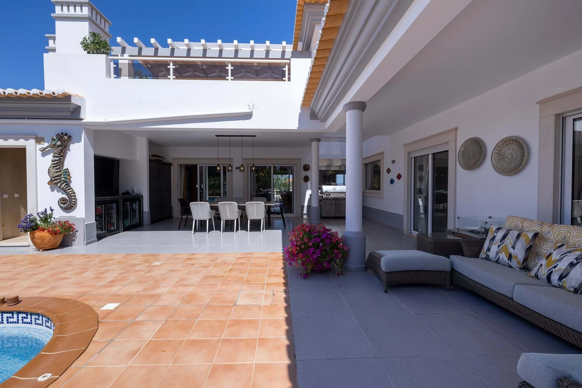 T5 Villa with pool in Ferrel 5mn from beach and Lagos centre – SunnySteve 49 White Mediterranean-style patio with tiled courtyard, outdoor dining table for six, and a built-in grill under a shaded area; potted flowers and a seahorse wall sculpture nearby.