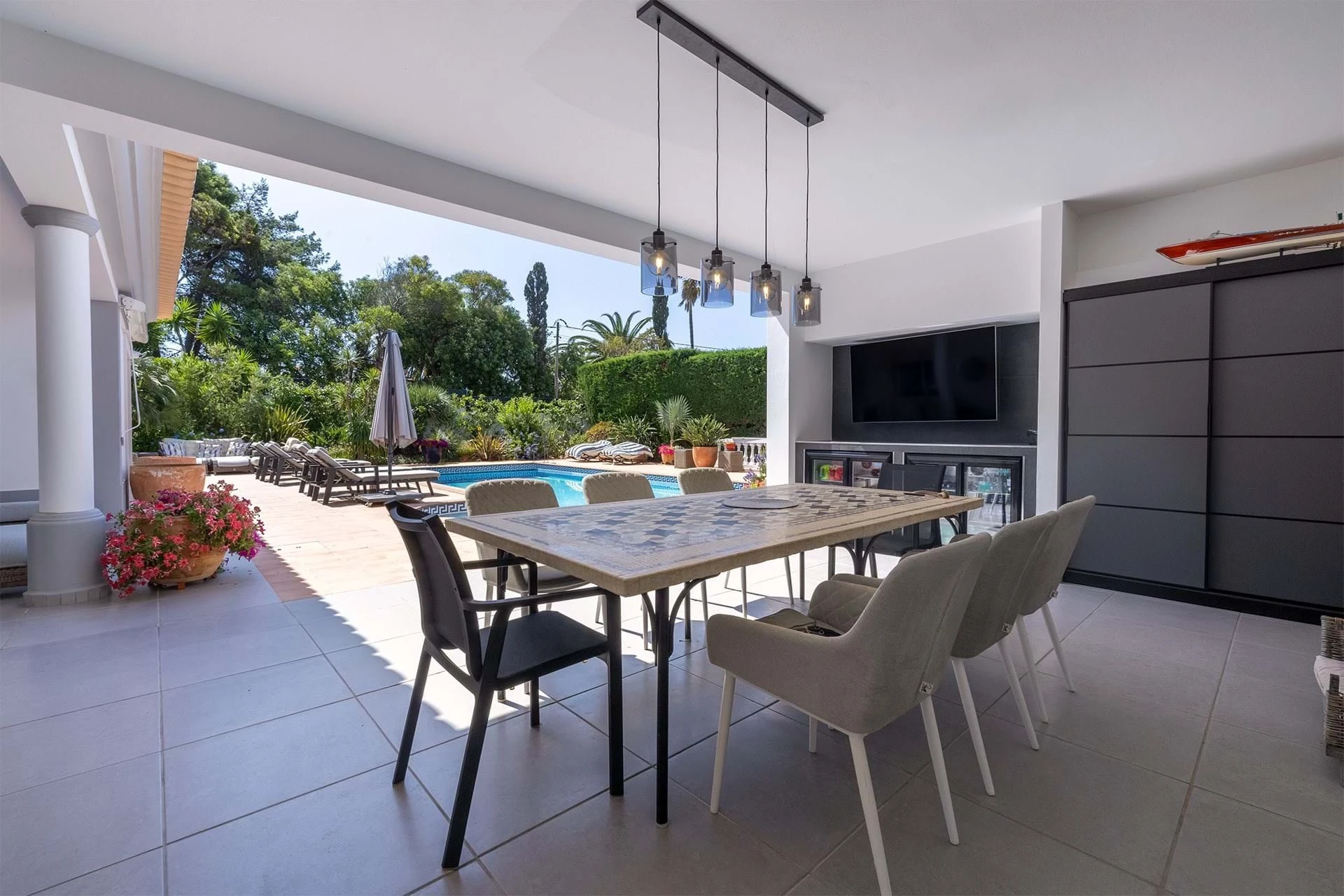 T5 Villa with pool in Ferrel 5mn from beach and Lagos centre – SunnySteve 5 Covered outdoor patio with a long dining table, beige chairs, and hanging pendant lights overlooking a pool and lush garden