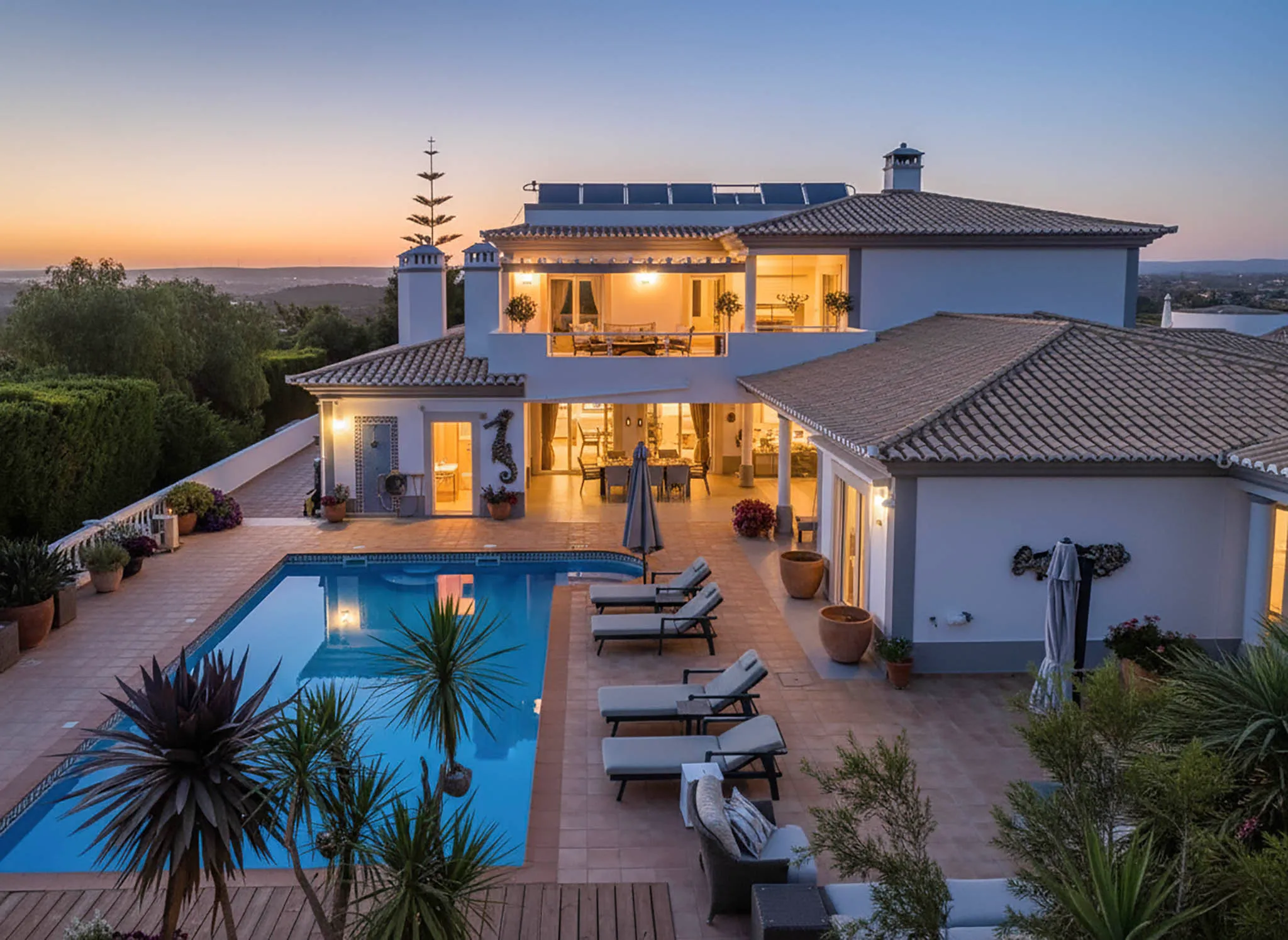 T5 Villa with pool in Ferrel 5mn from beach and Lagos centre – SunnySteve 56 Evening view of a modern villa with a rectangular pool, lounge area, and potted plants, illuminated as the sun sets on the horizon.
