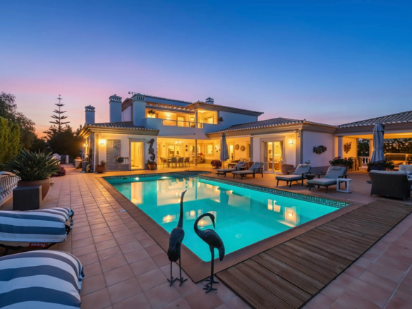 Luxurious white villa with a lit interior, outdoor seating, and a turquoise pool at dusk.