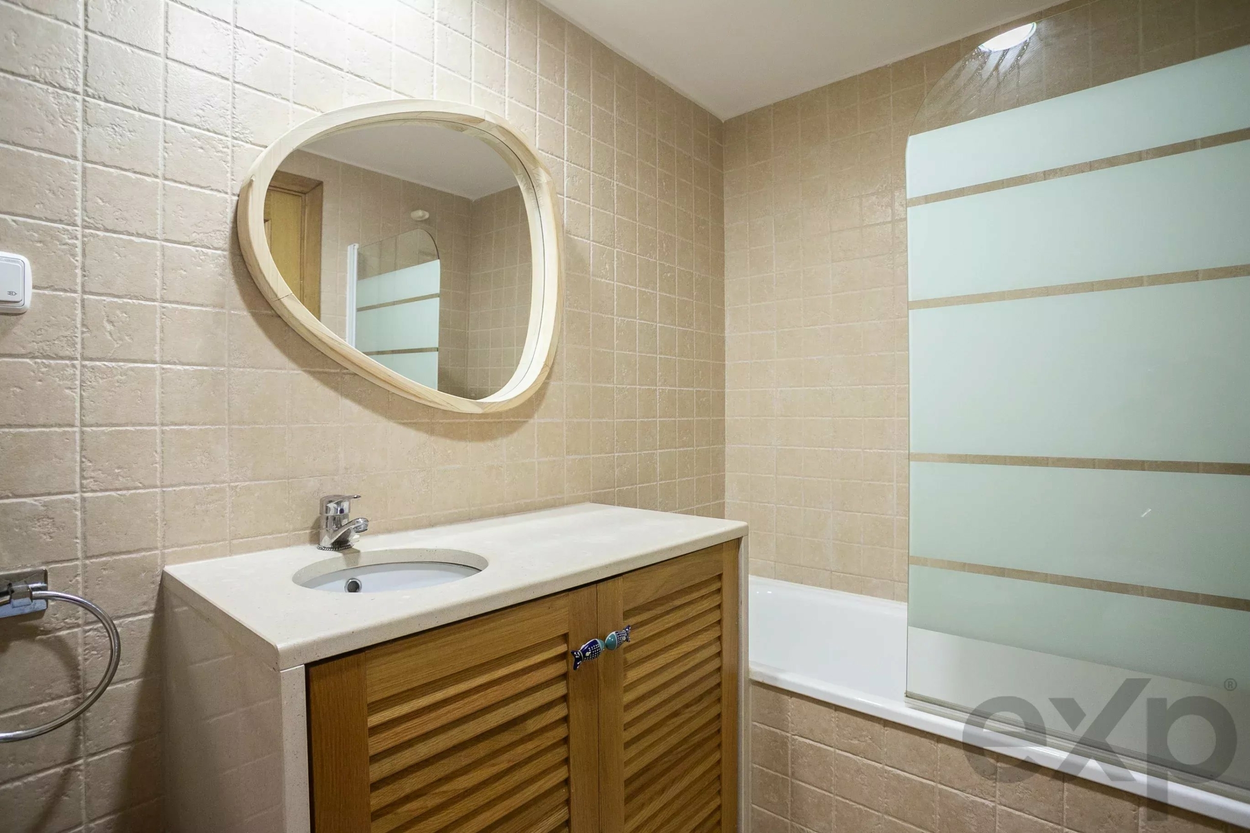Round wall mirror above a beige countertop sink with a wooden slatted vanity cabinet in a tiled bathroom.