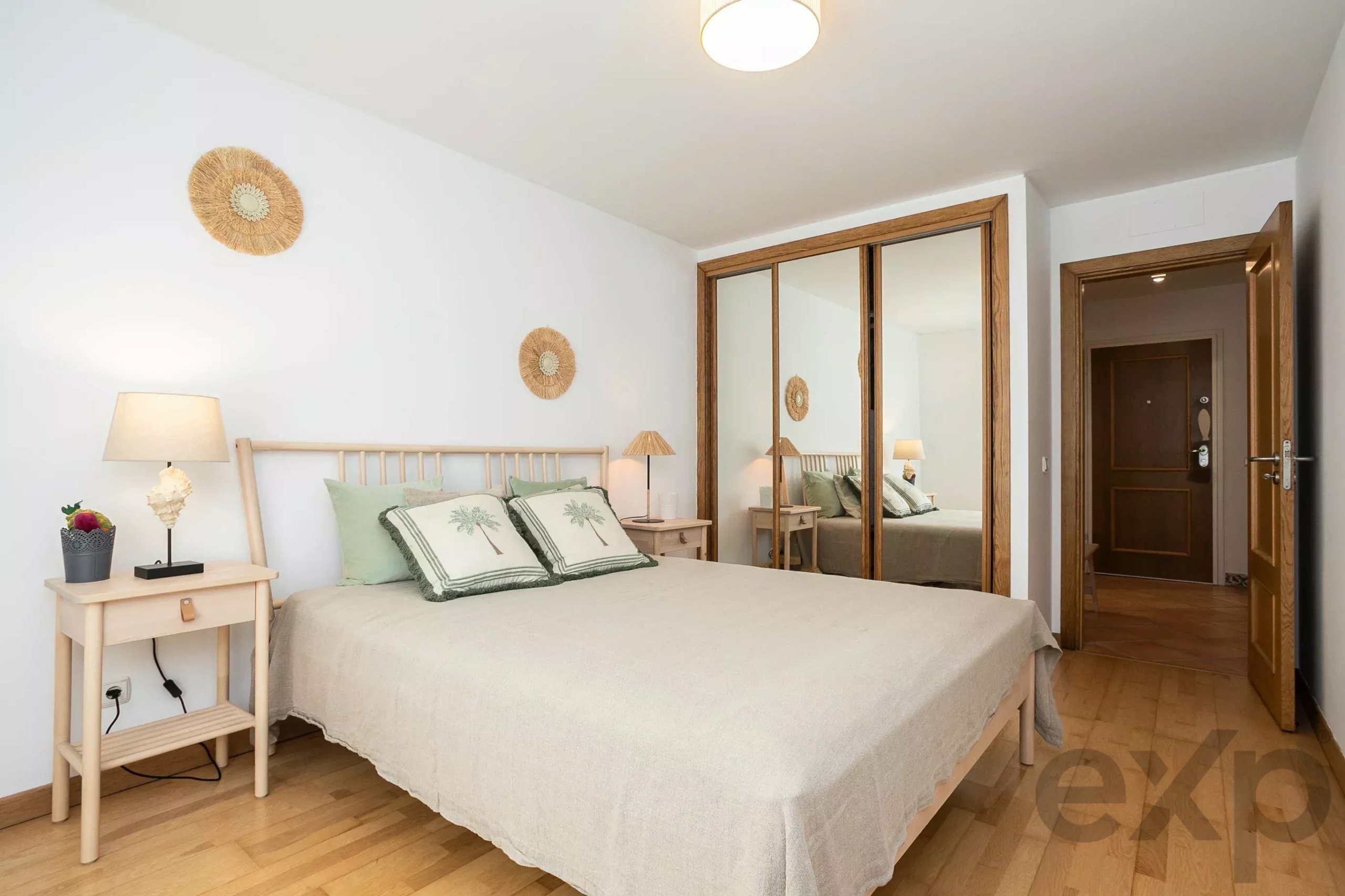 Bright bedroom with a beige duvet, two wooden nightstands, and warm lamps; a large mirrored wardrobe reflects the bed. Open door to a hallway visible on the right.