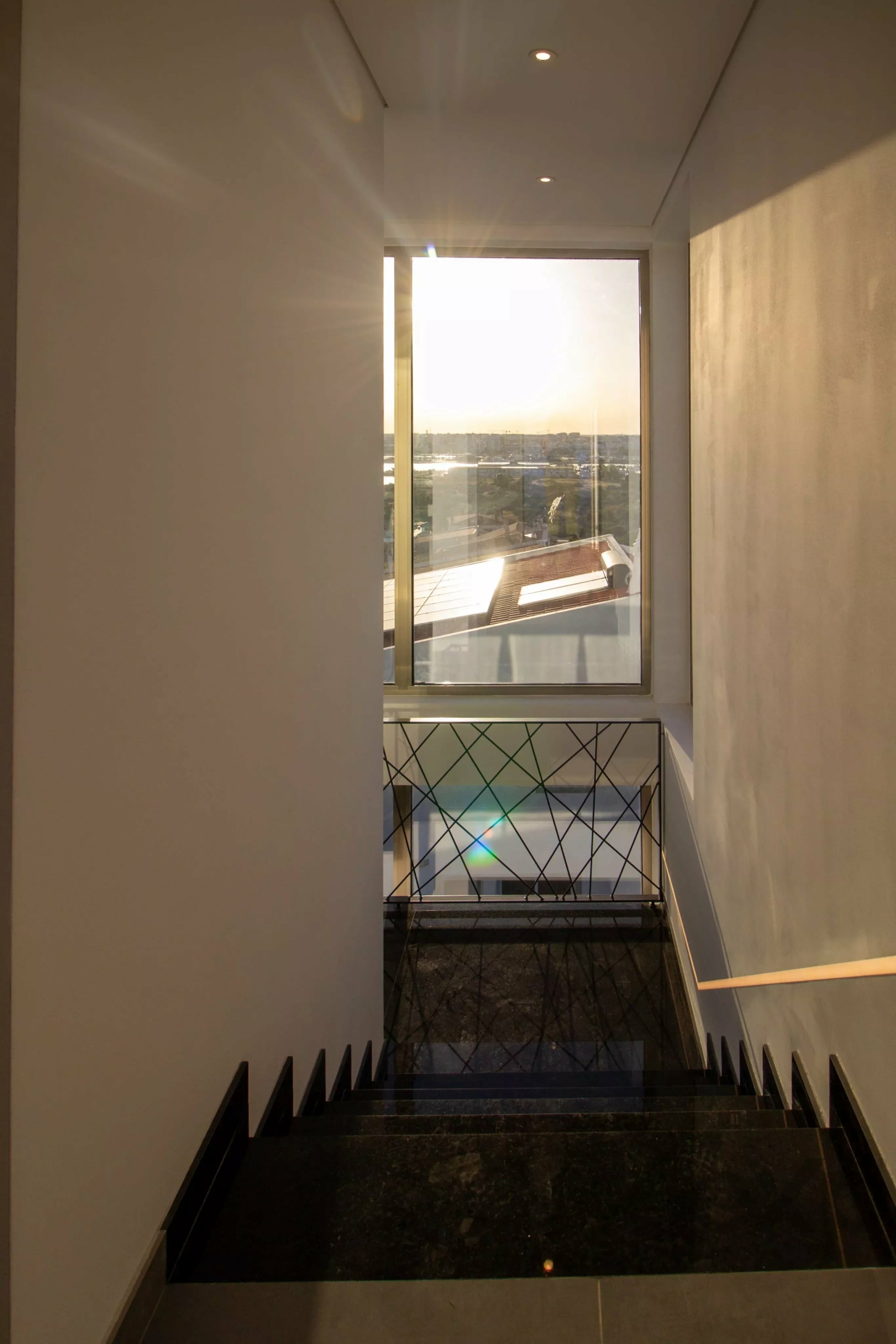 View of sunlit cityscape through a large window at the end of a narrow hallway, seen from a staircase landing.