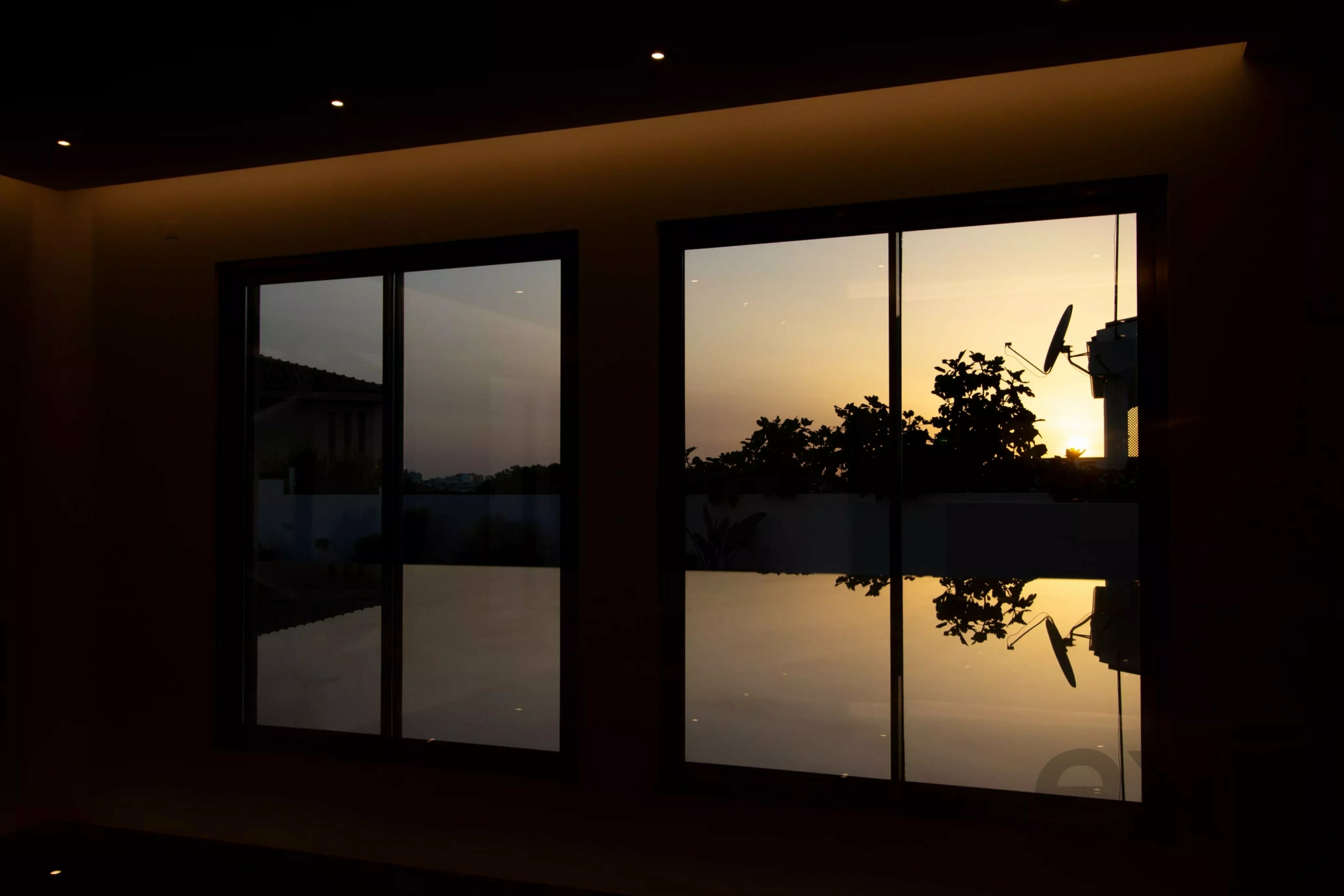 Dim interior with three large glass windows showing a sunset, silhouetted trees, and a satellite dish outside.