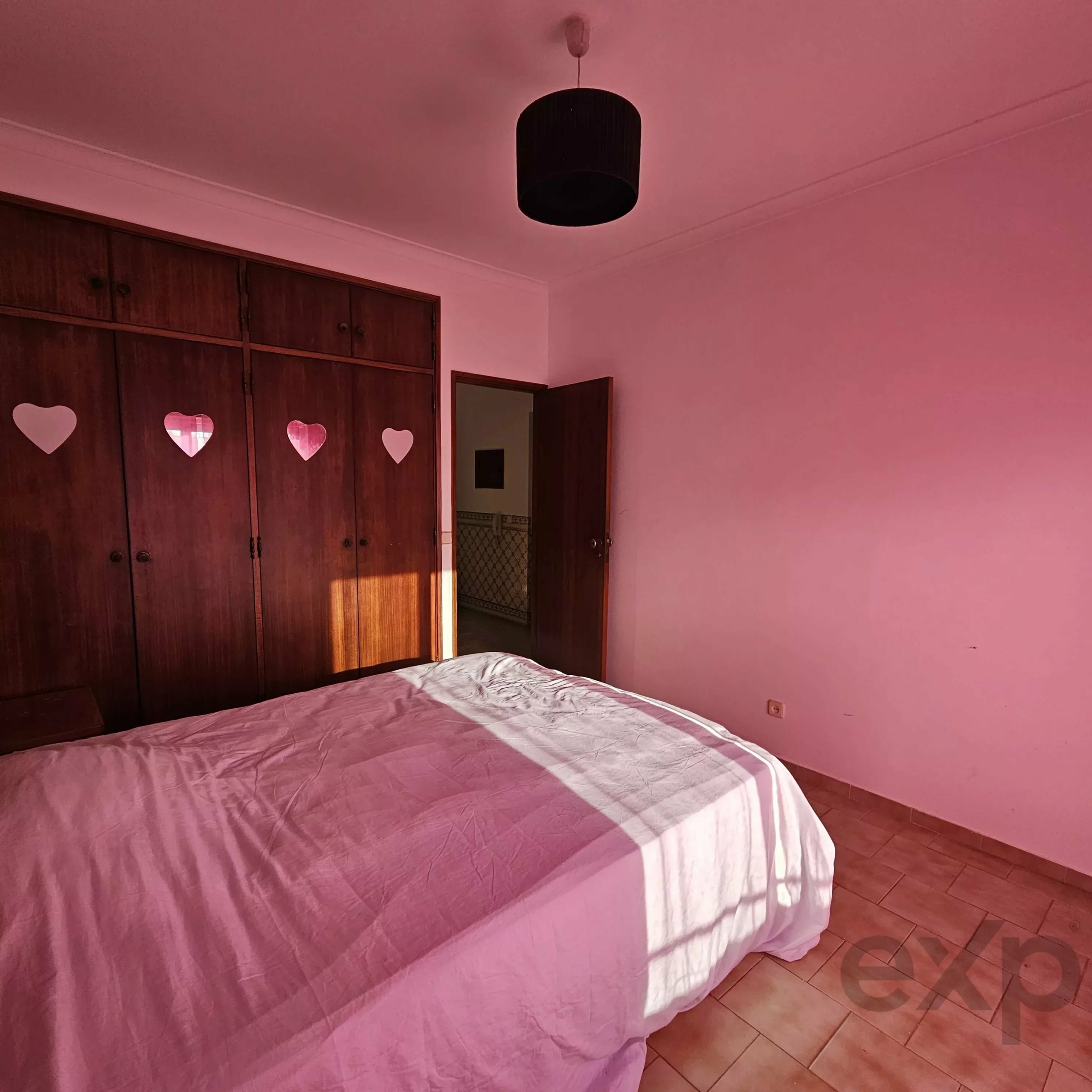 Bedroom with pink lighting, two single beds covered in pink quilts, dark wooden wardrobes, and a black cylindrical ceiling light.