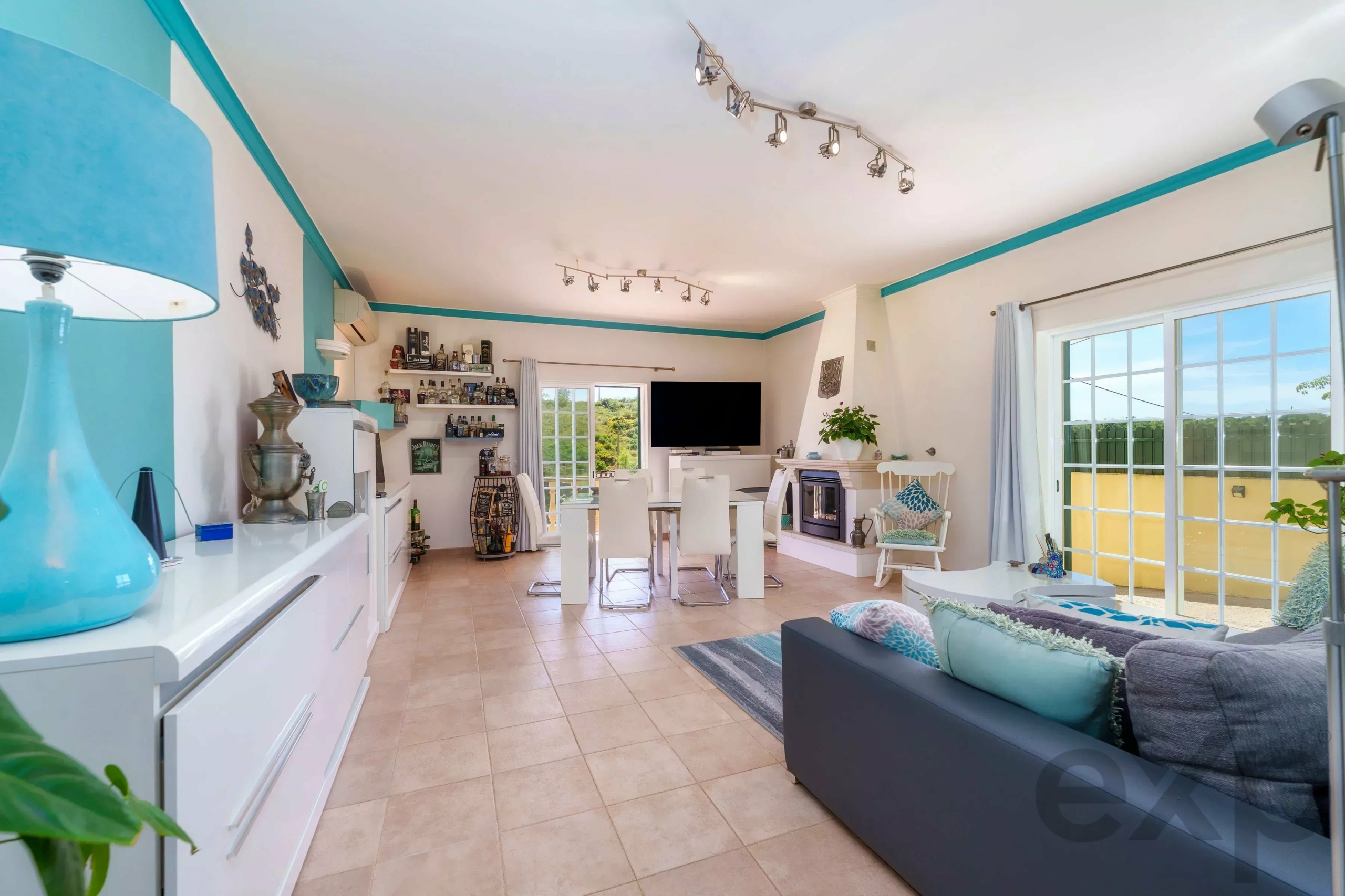 Bright open-plan living room with turquoise trim, white furniture, and large glass doors to a sunny outdoor area. Track lighting runs along the ceiling.