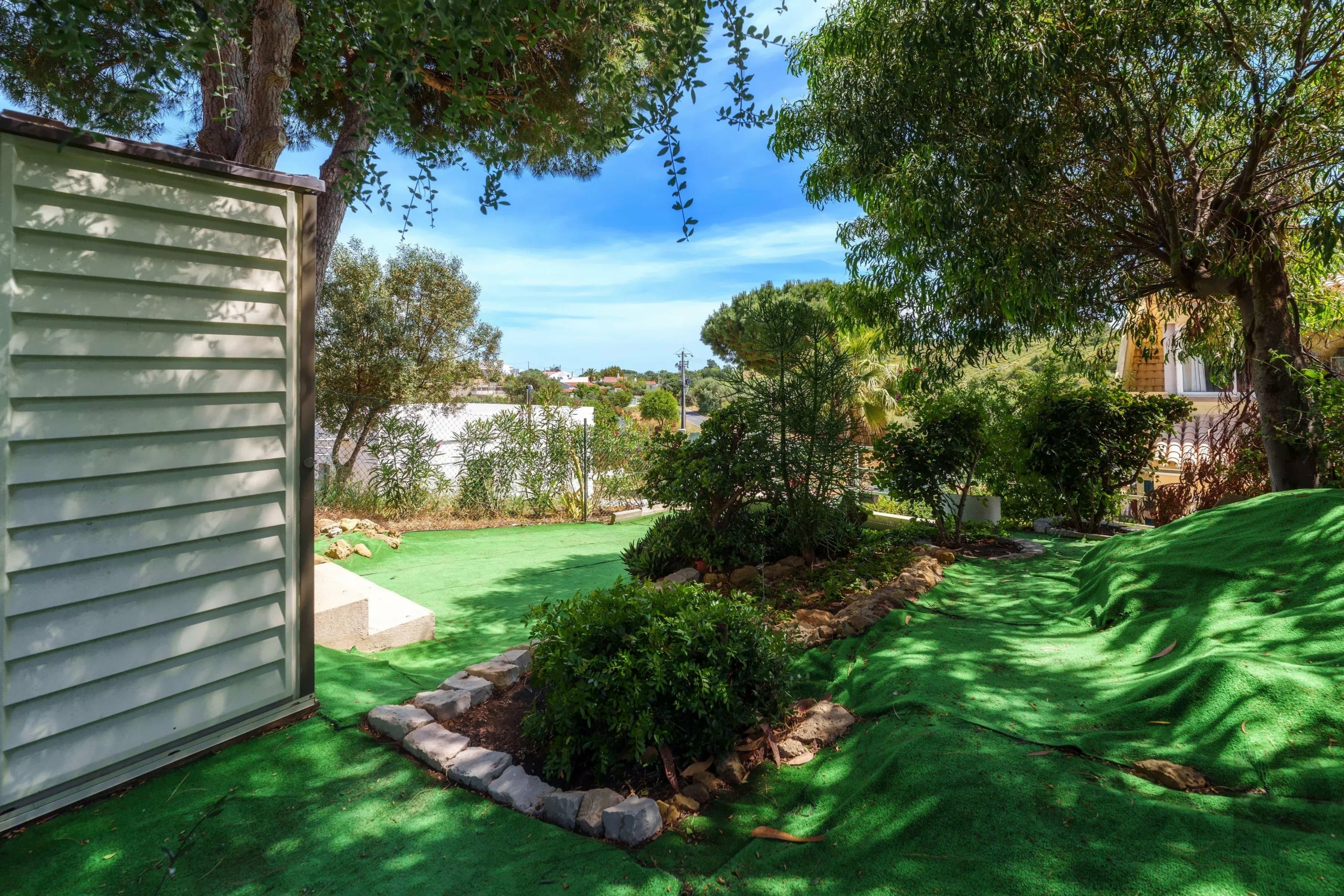 Green artificial turf lawn with stone edging and shrubs, seen beside a light green shed under leafy trees against a blue sky.
