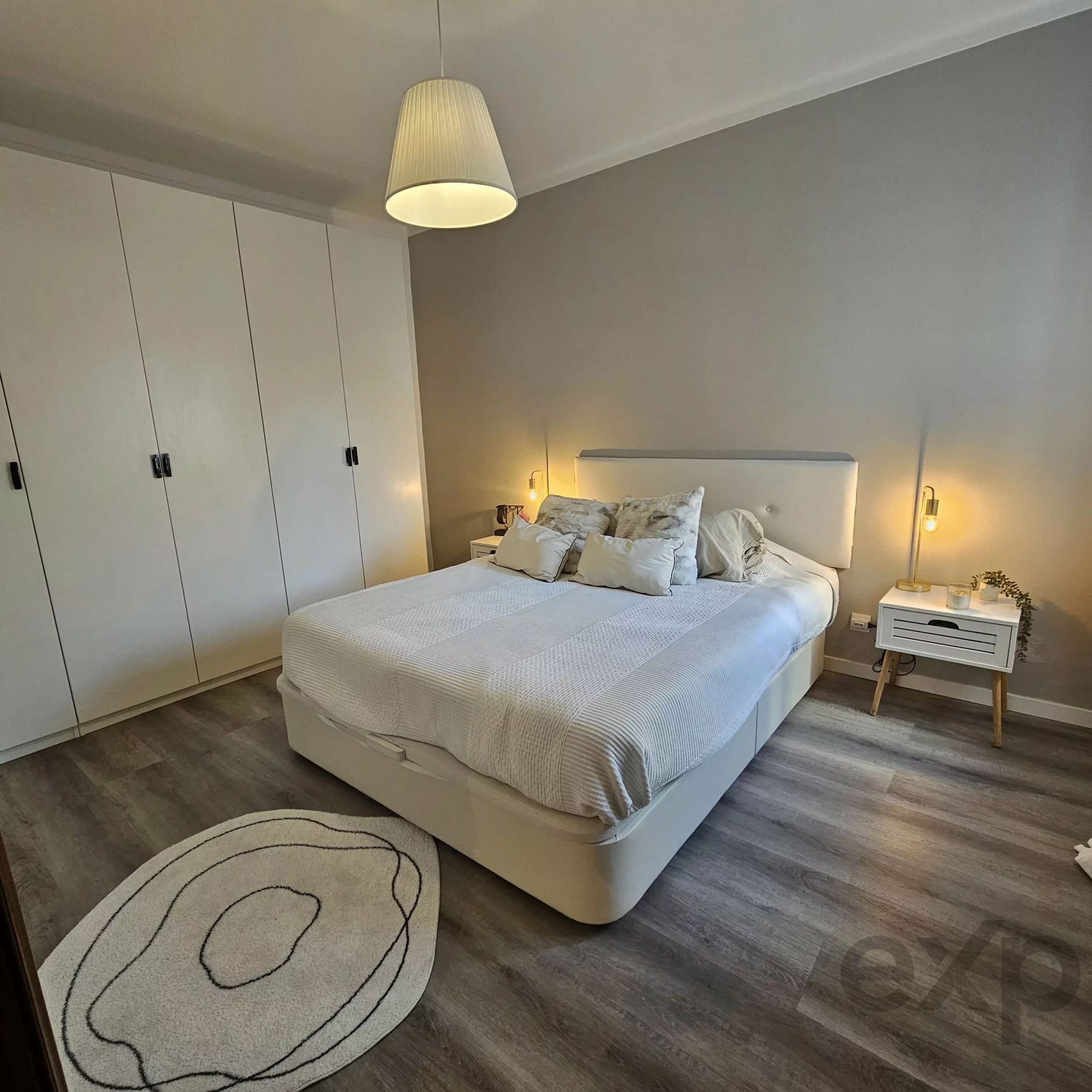 Modern minimalist bedroom with a large white bed, matching nightstands, built-in white wardrobes along the left wall, and a hanging pendant light above.