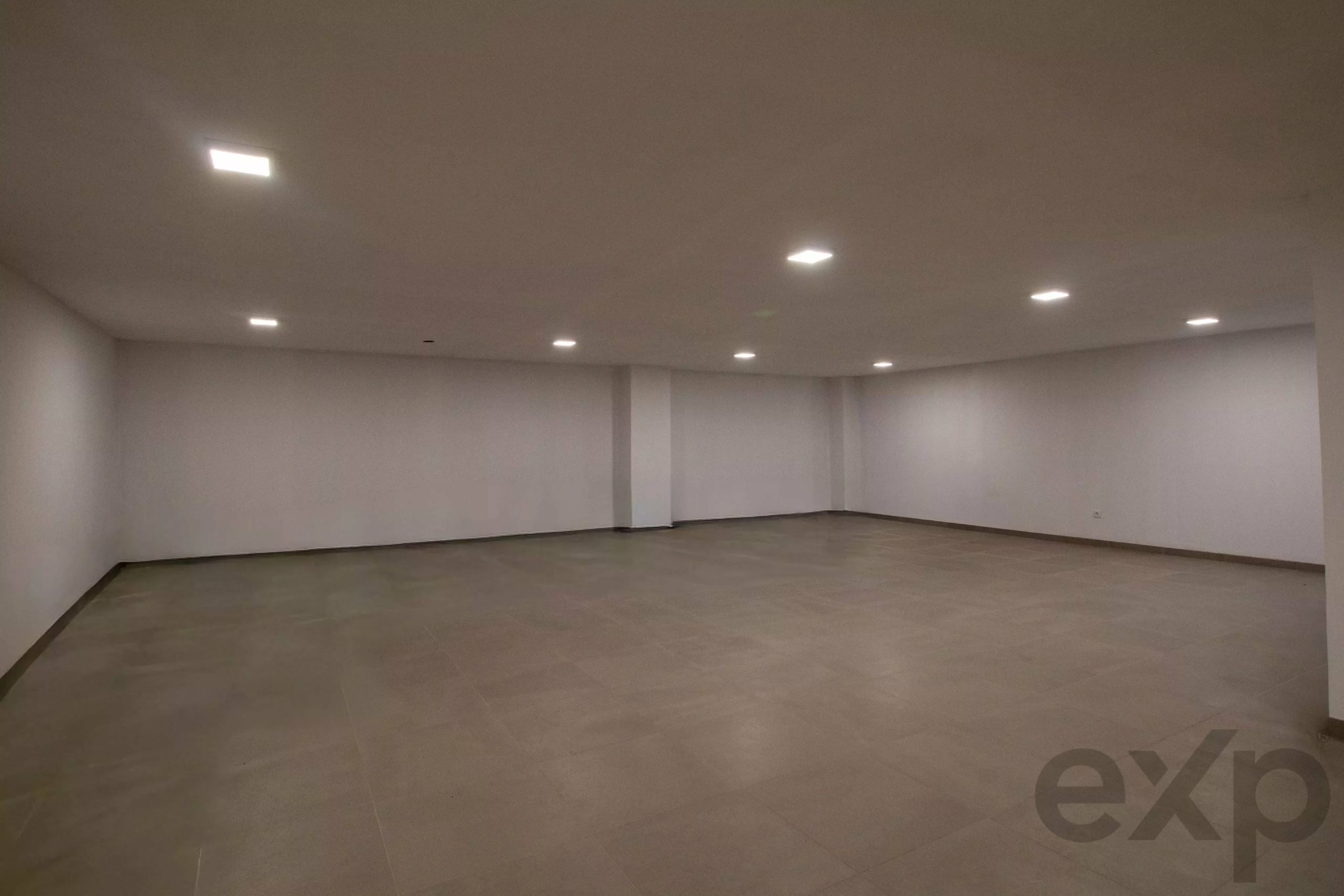 Empty white-walled exhibition space with tiled floor and multiple recessed ceiling lights.