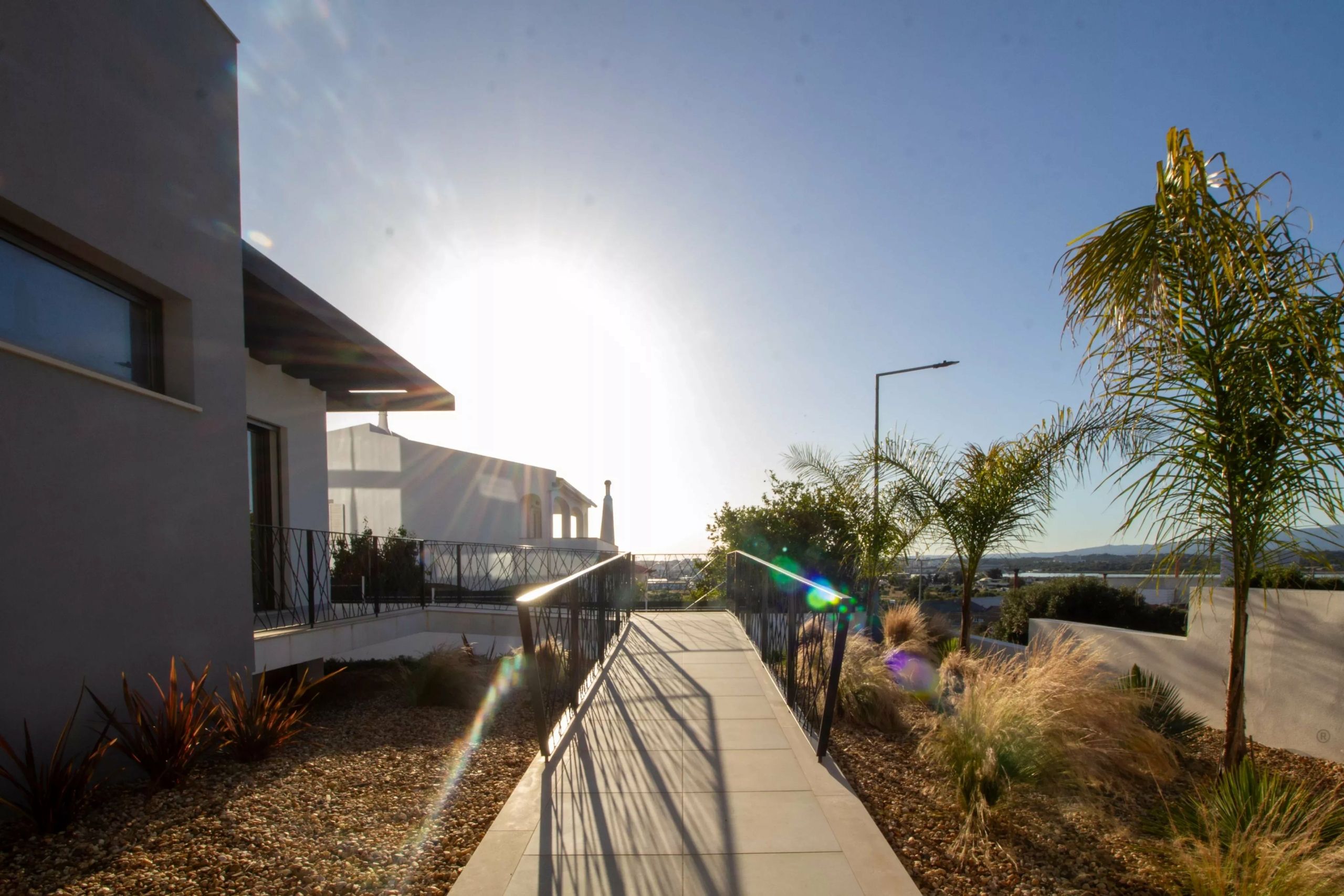 Sunlit modern house exterior with a walkway, railing, and palm trees under a clear blue sky.