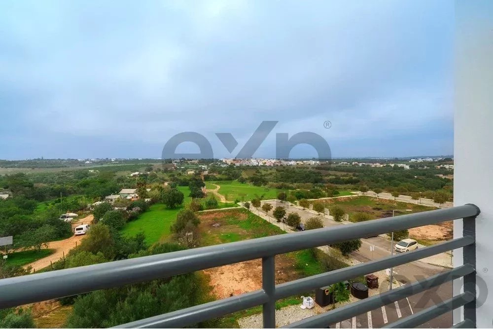 Balcony railing overlooks a green suburban landscape with trees, fields, and distant houses under a cloudy sky.