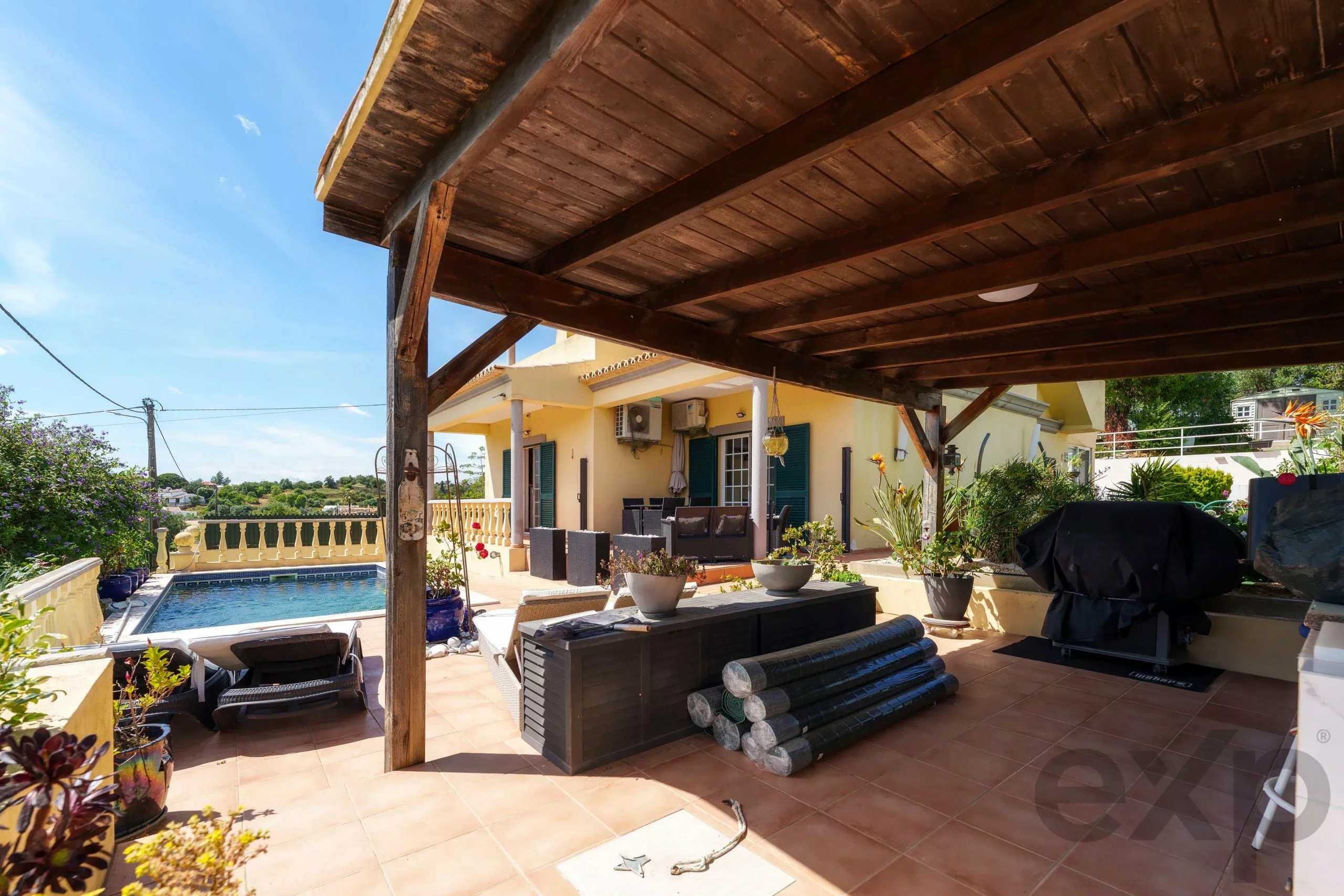 Patio beneath a wooden pergola with outdoor seating, a rectangular pool, and a yellow house under a clear blue sky.