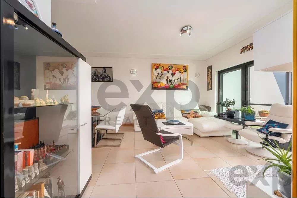 Bright modern living room with a white sectional sofa, black lounge chair, glass coffee table, and potted plants by large windows.