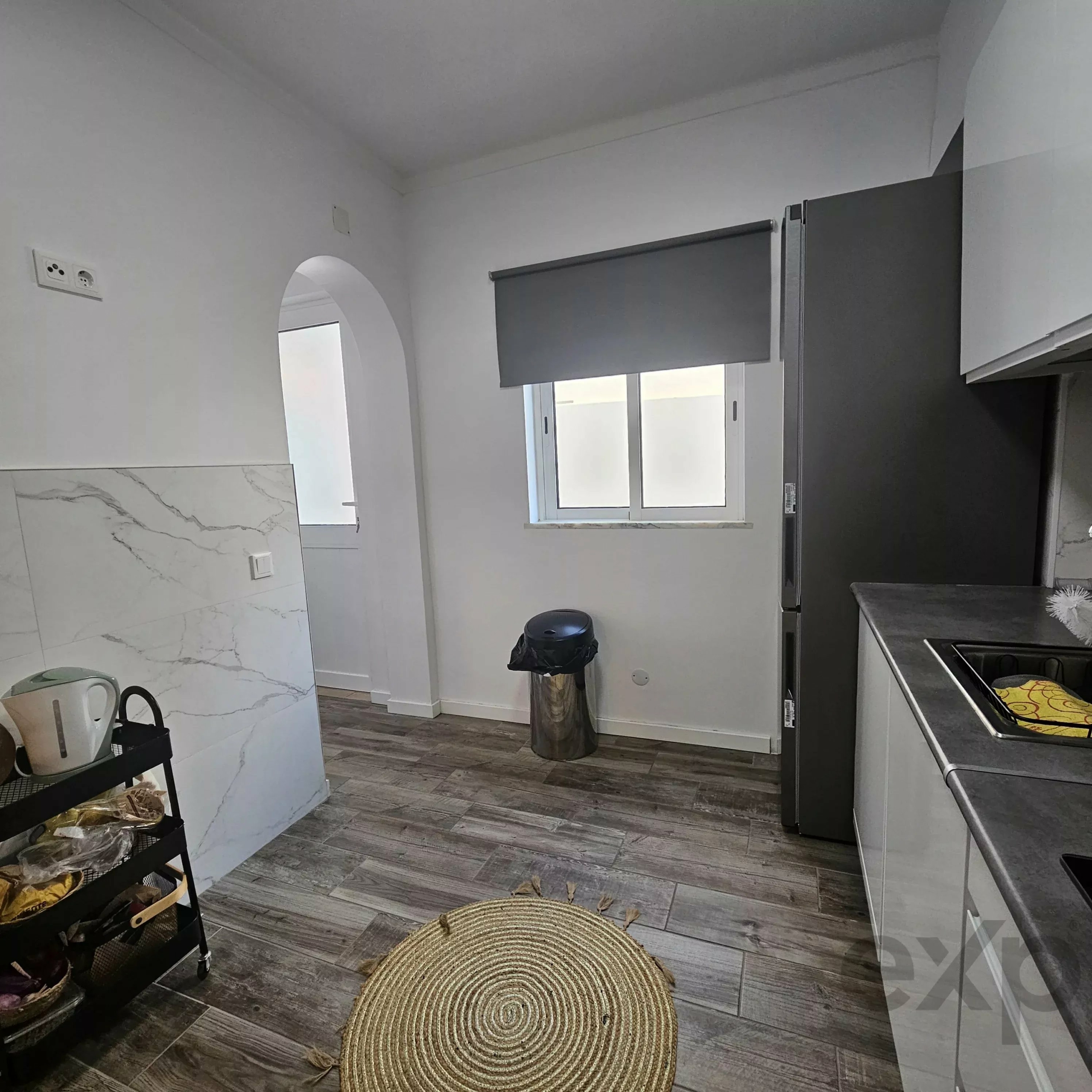 Modern kitchen with gray cabinets, window with roller shade, marble-tile wall on left, and a round woven rug on wood floor in the center.