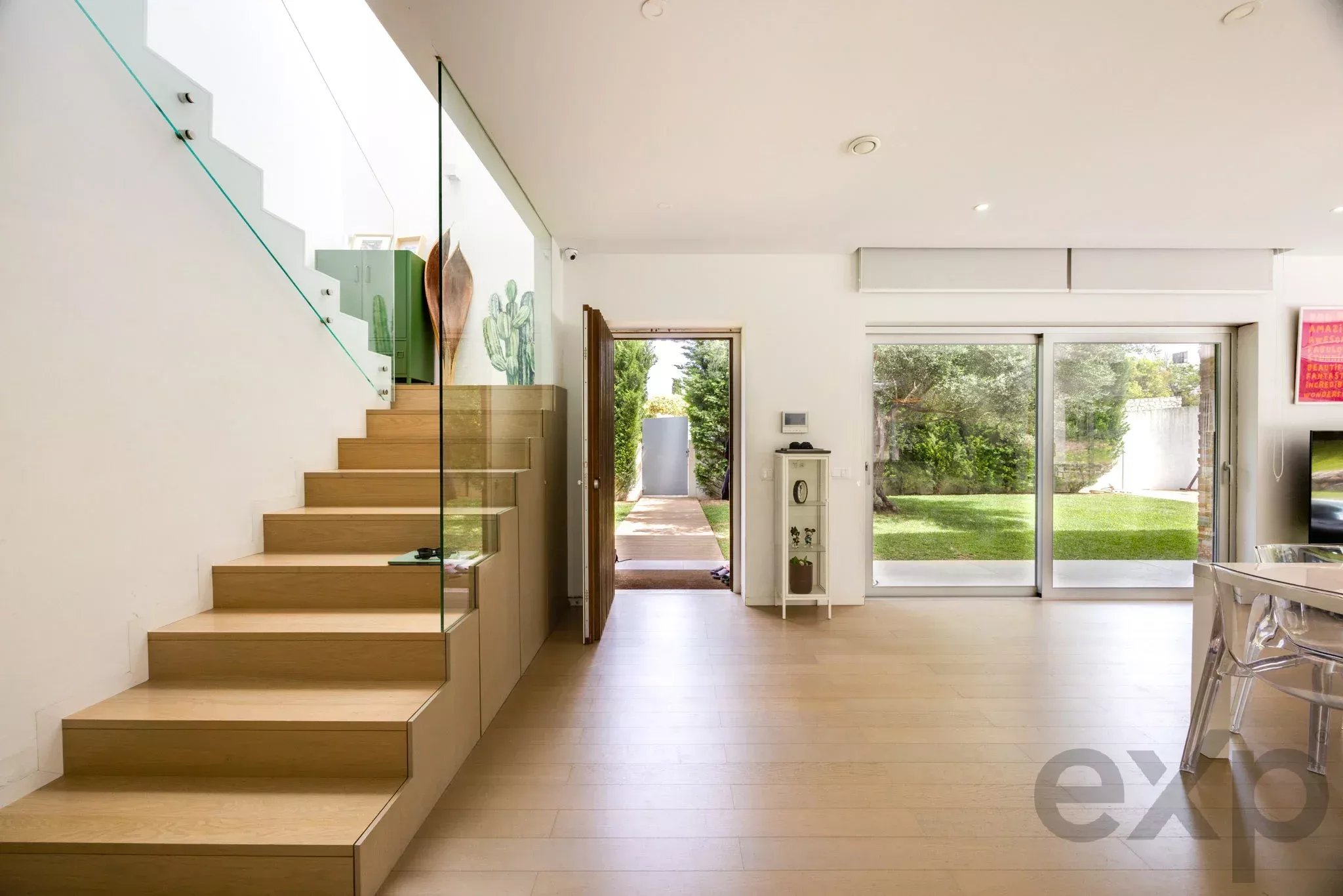 Bright modern living area with a wooden staircase and glass railing on the left, open doorway to a garden, and large sliding glass doors to the yard on the right.