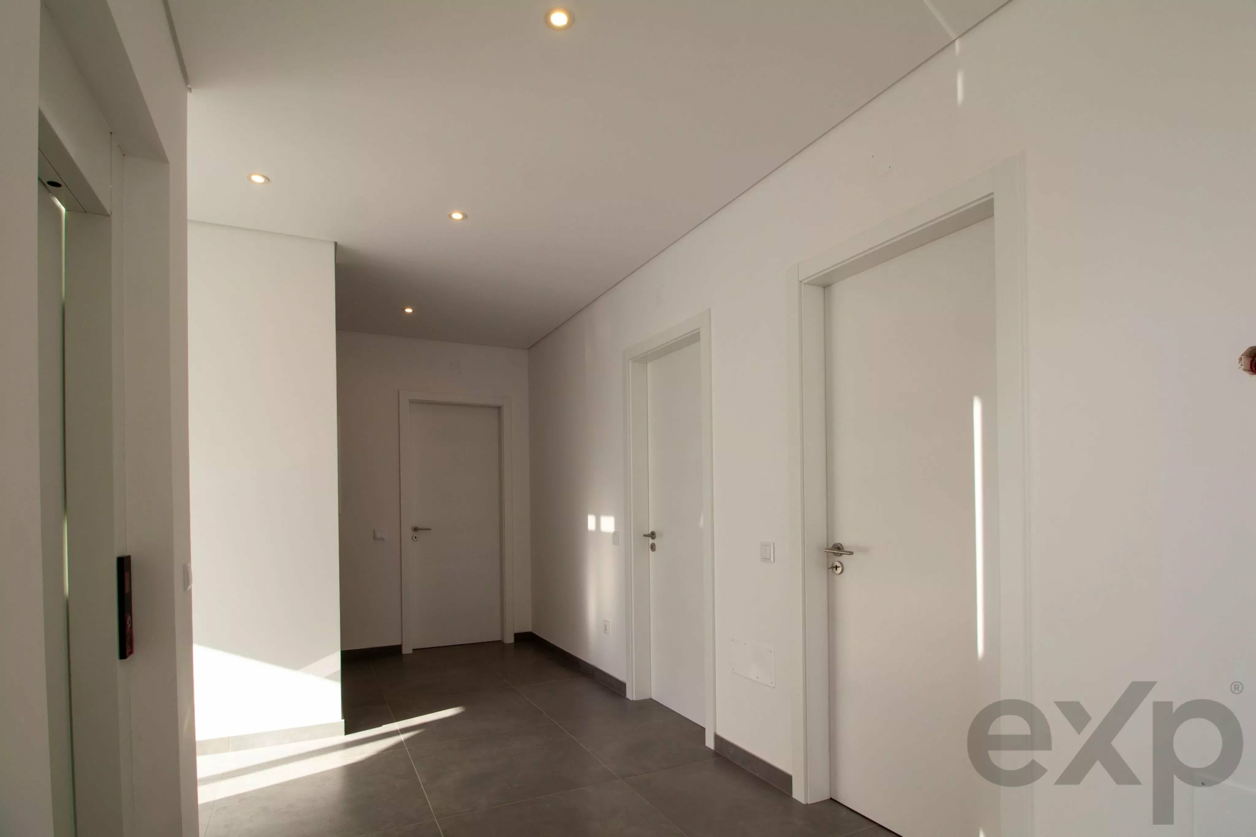 White modern hallway with closed white doors along the right wall and recessed ceiling lights, gray tile floor, and natural light patterns on the floor.