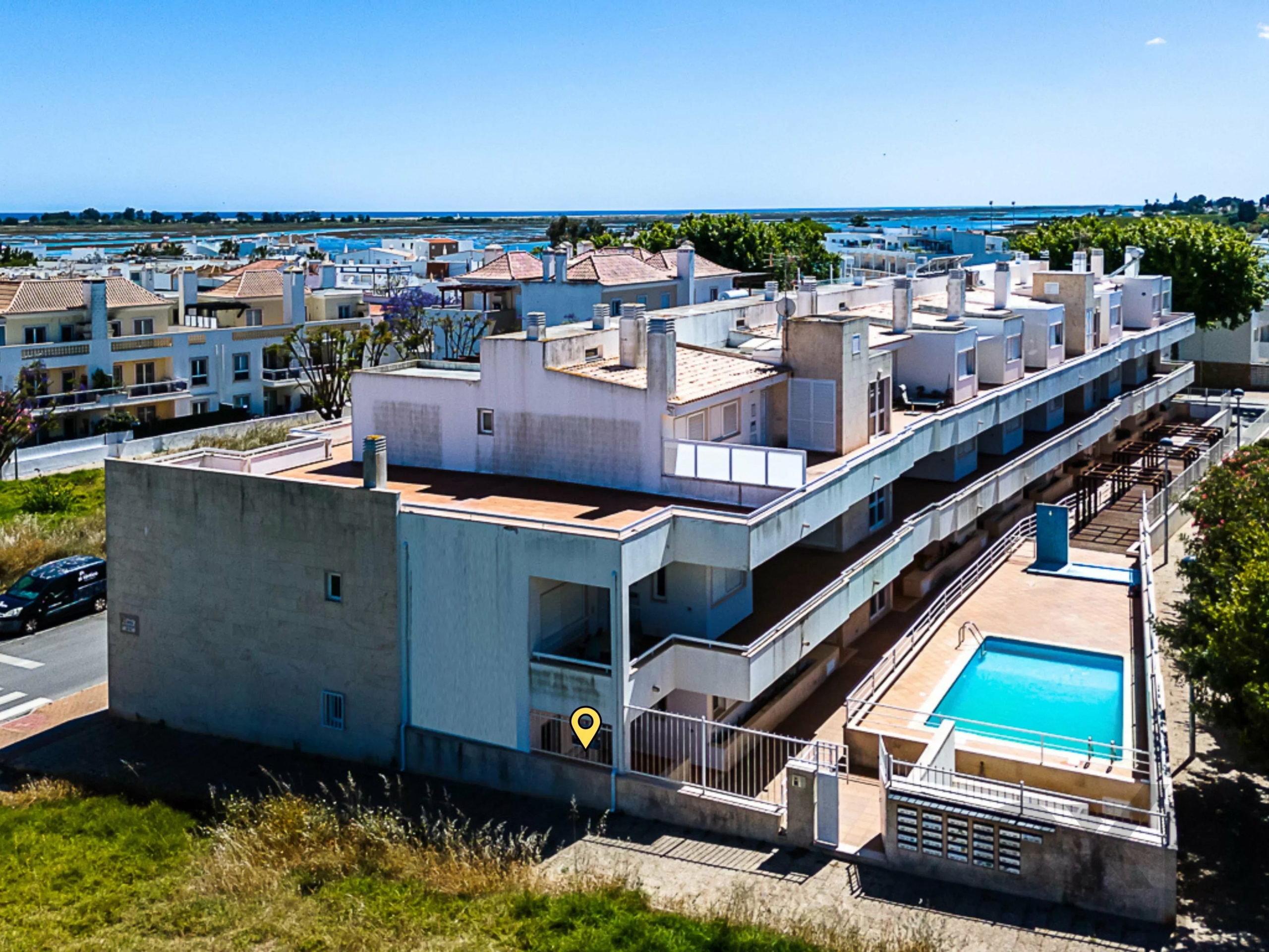 Aerial view of white apartment buildings with balconies, a blue rectangular pool in the foreground, and the sea on the horizon.