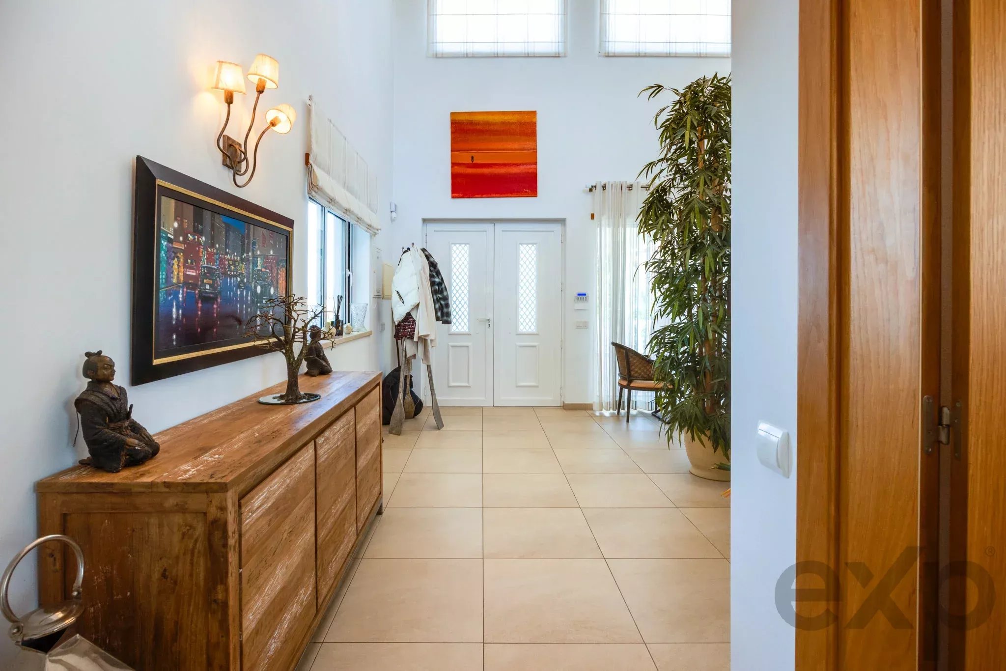 Cozy hallway with a rustic wooden sideboard, bronze statues, and wall art, leading to a white door and a tall plant on the right.