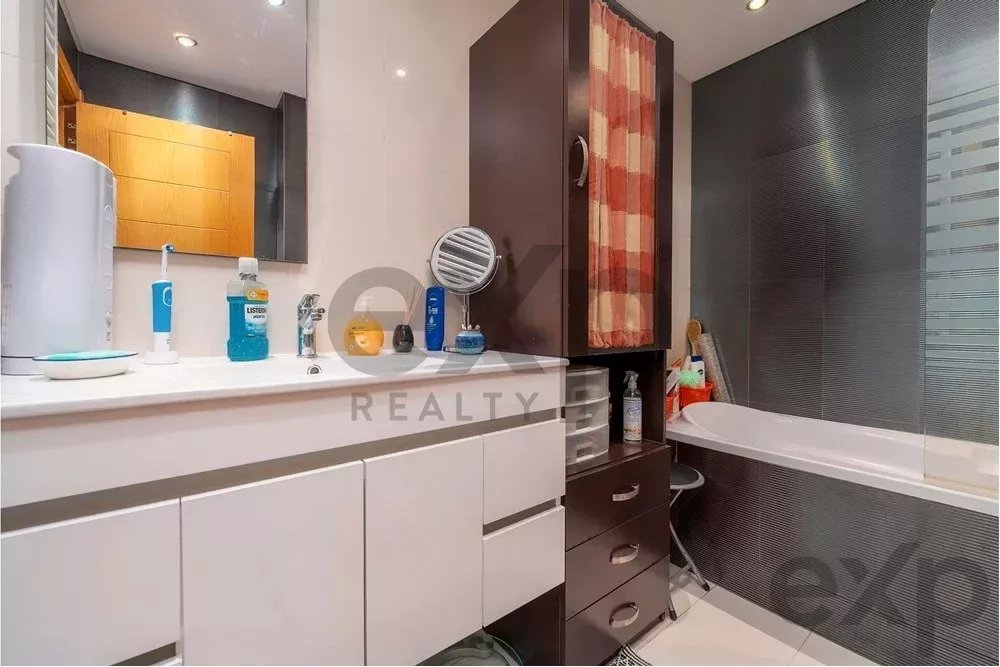 Modern bathroom with white vanity and counter, mirror, and a tall dark storage cabinet with a pink striped curtain; bathtub along dark tile wall.