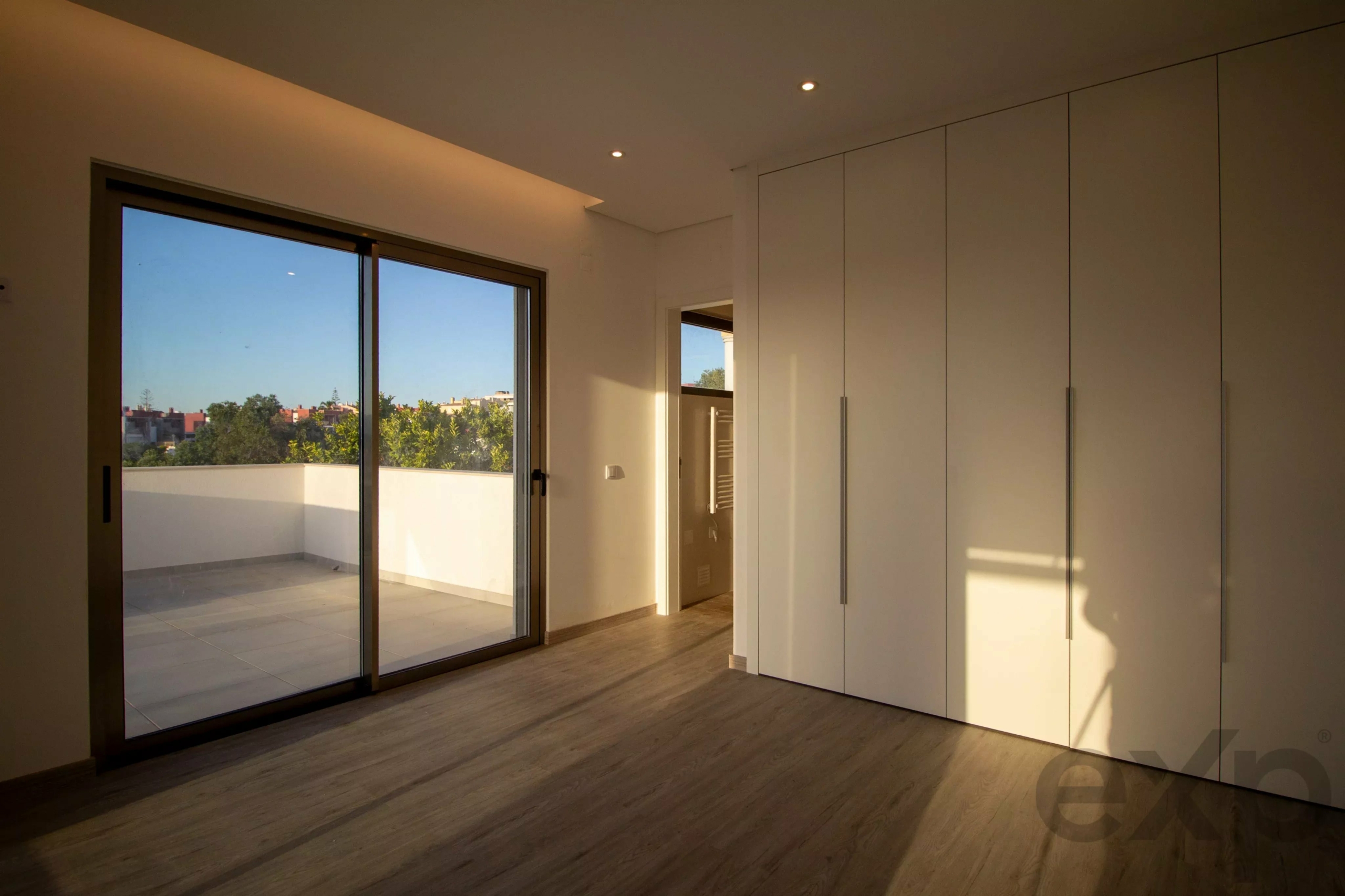 Bright modern room with white floor-to-ceiling wardrobes on the right and a large glass sliding door to a balcony, trees and buildings visible outside.