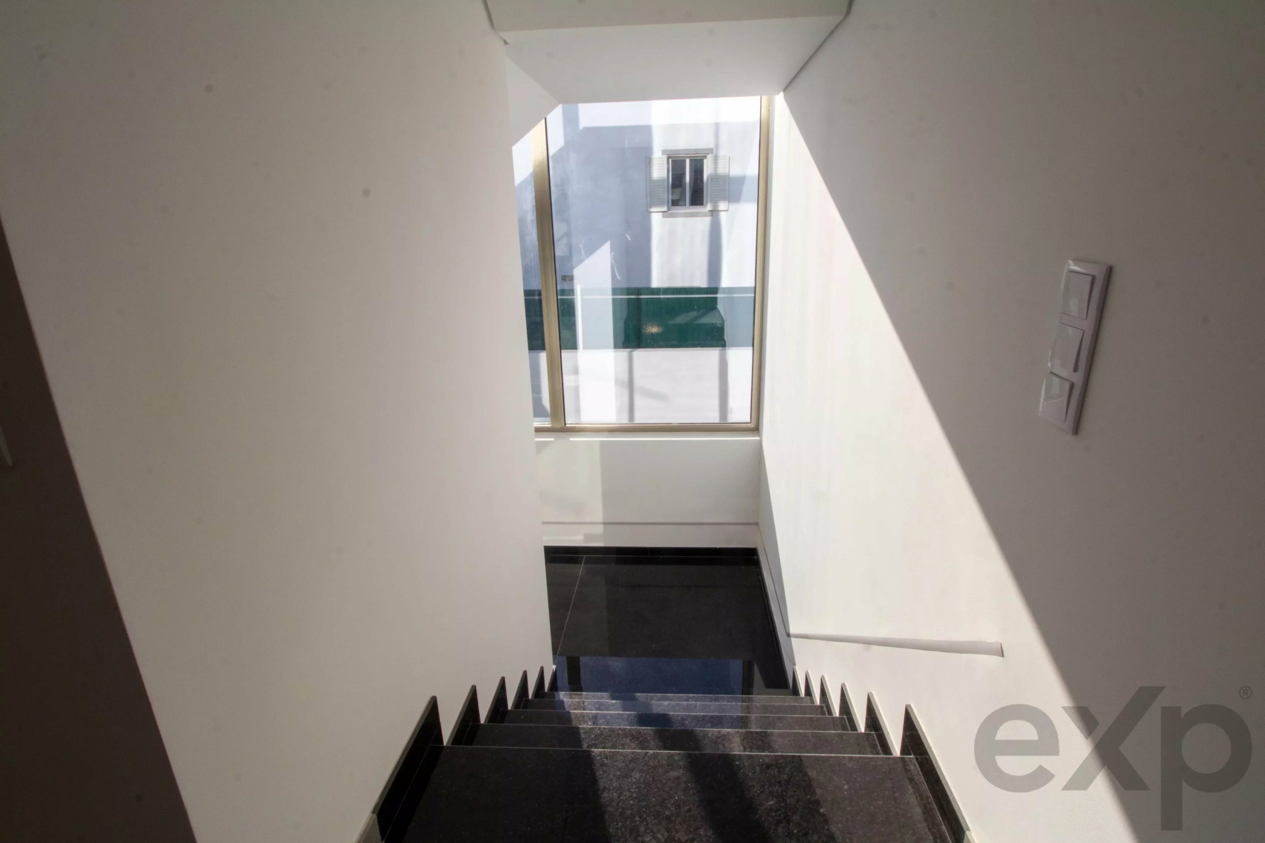 Downward view of a modern staircase with black steps and white walls, ending at a glass door with a large window outside, letting in bright light.