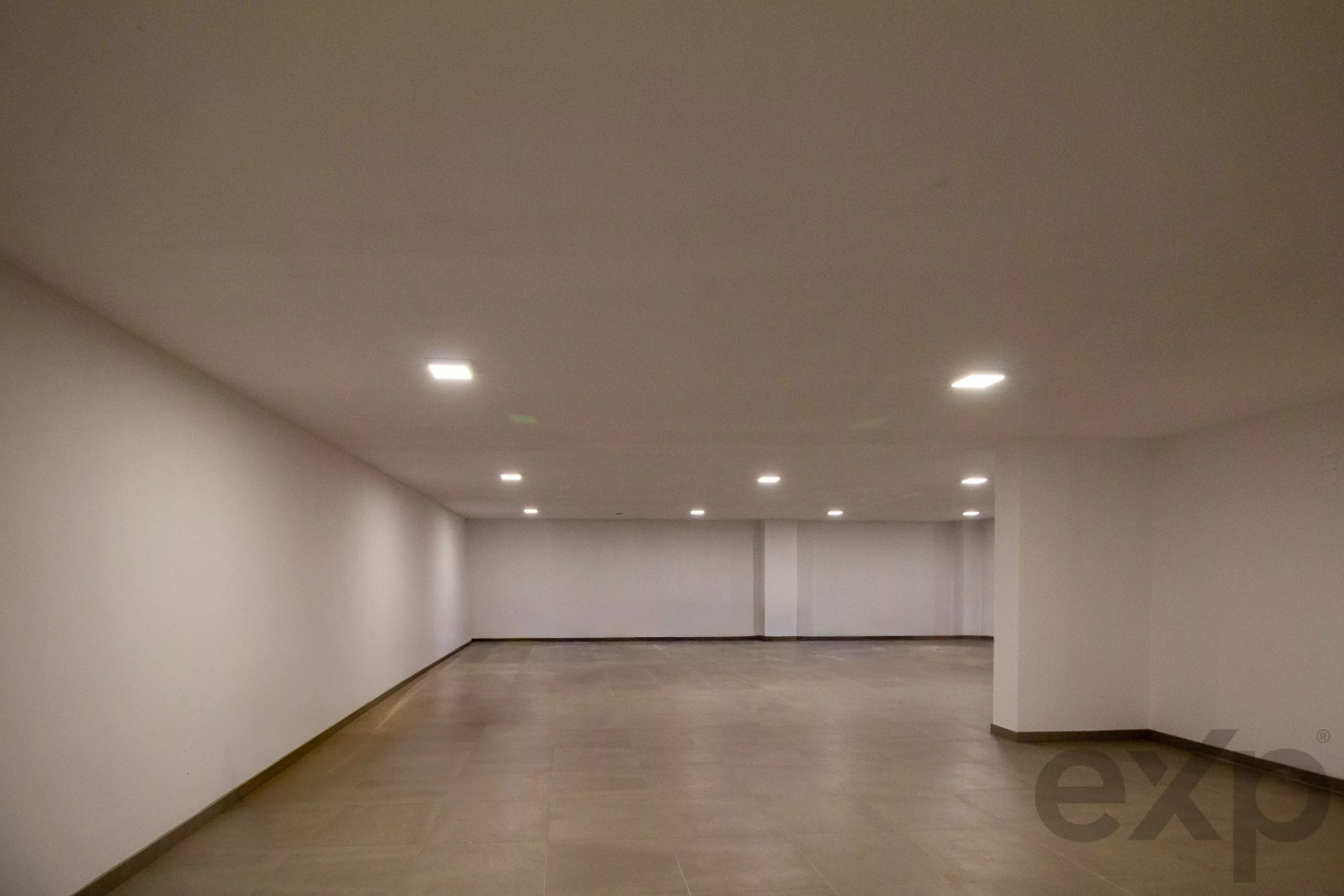 Long empty hallway with white walls, tiled floor, and recessed ceiling lights receding into the distance.