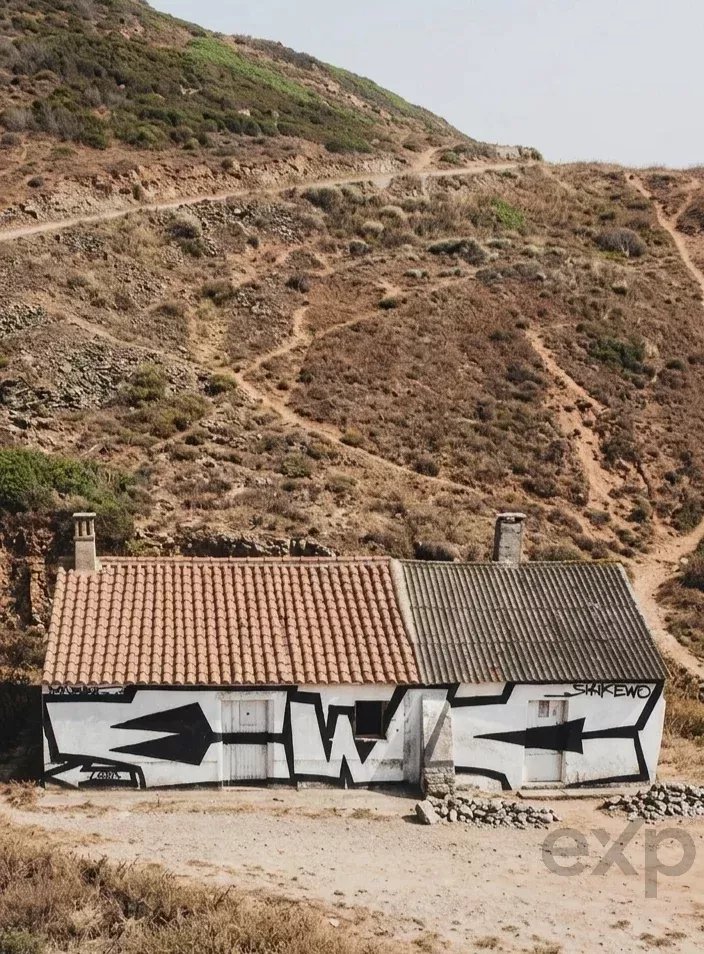 Graffiti-covered single-story house on a dry hillside, with a red-tiled roof on the left and a metal roof on the right.
