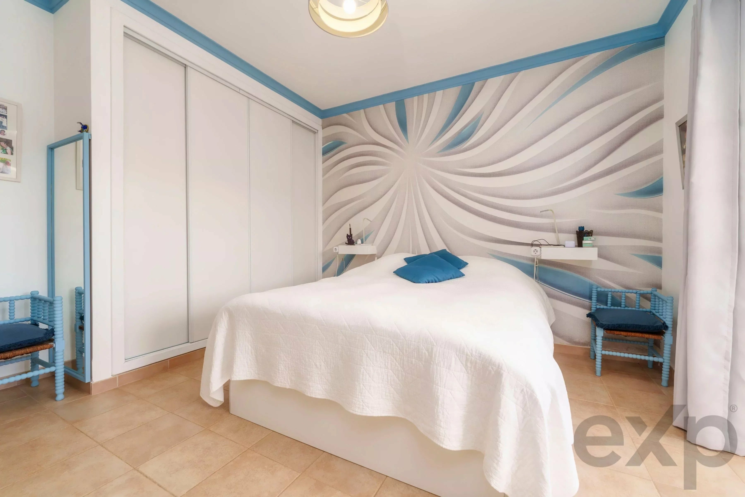 Bedroom with white and blue palette; bed with white quilt and blue cushions against a swirling grey-blue mural wall and white wardrobes nearby.