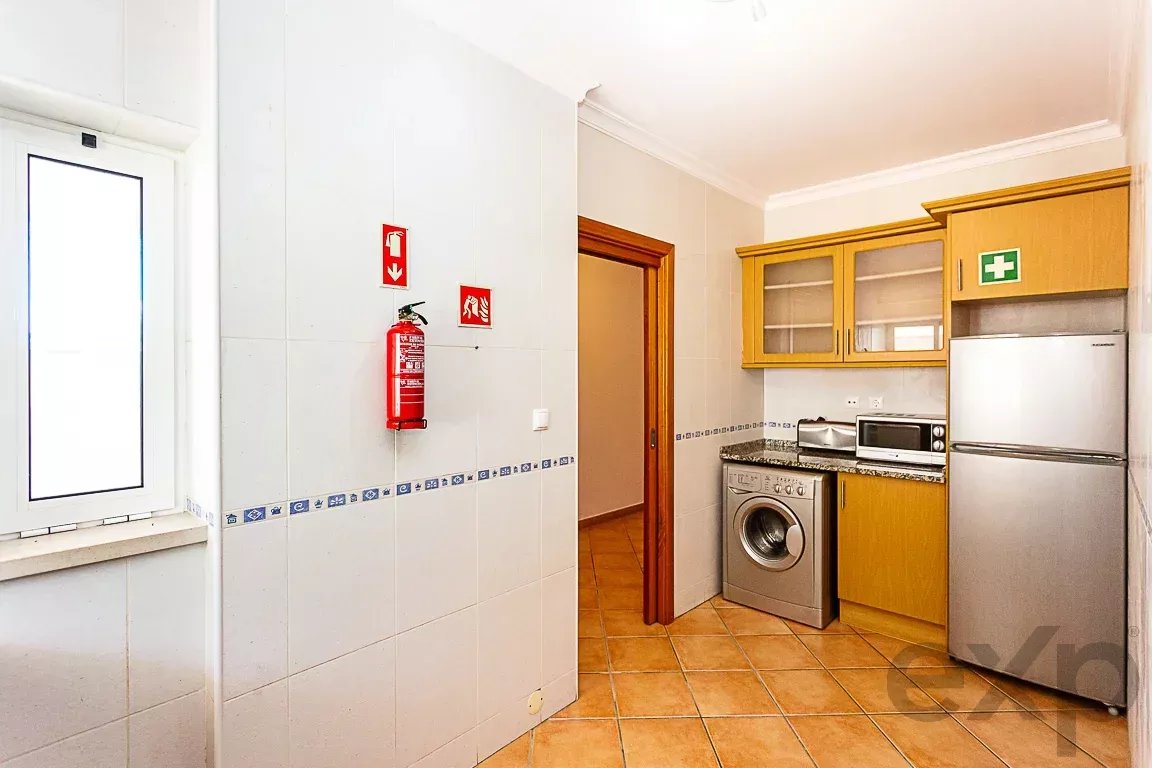 Bright kitchen with white tiled walls, a red fire extinguisher on the wall, a doorway to the left, and stainless steel fridge, washing machine, microwave, and wooden cabinets nearby.