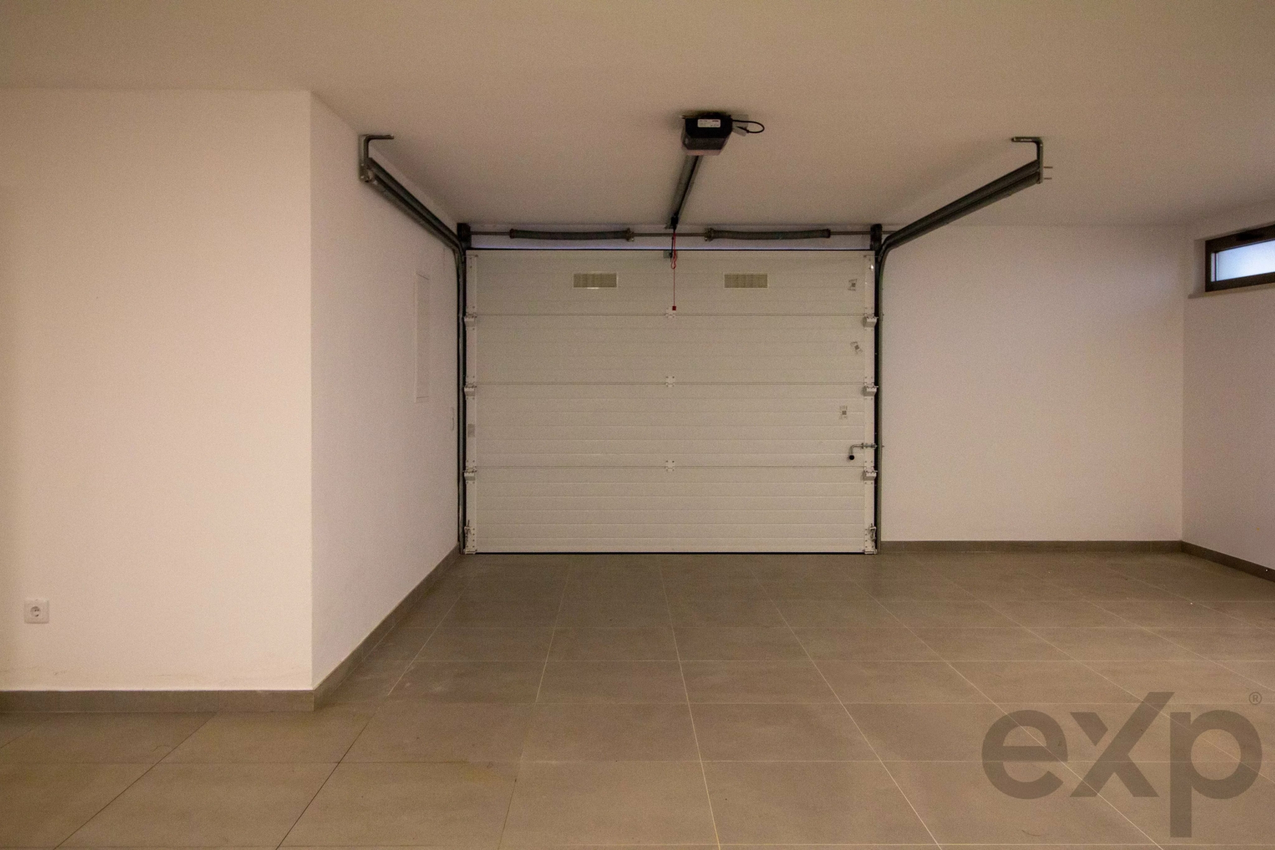 Garage interior with a closed white sectional door, ceiling-mounted opener, and tiled floor.