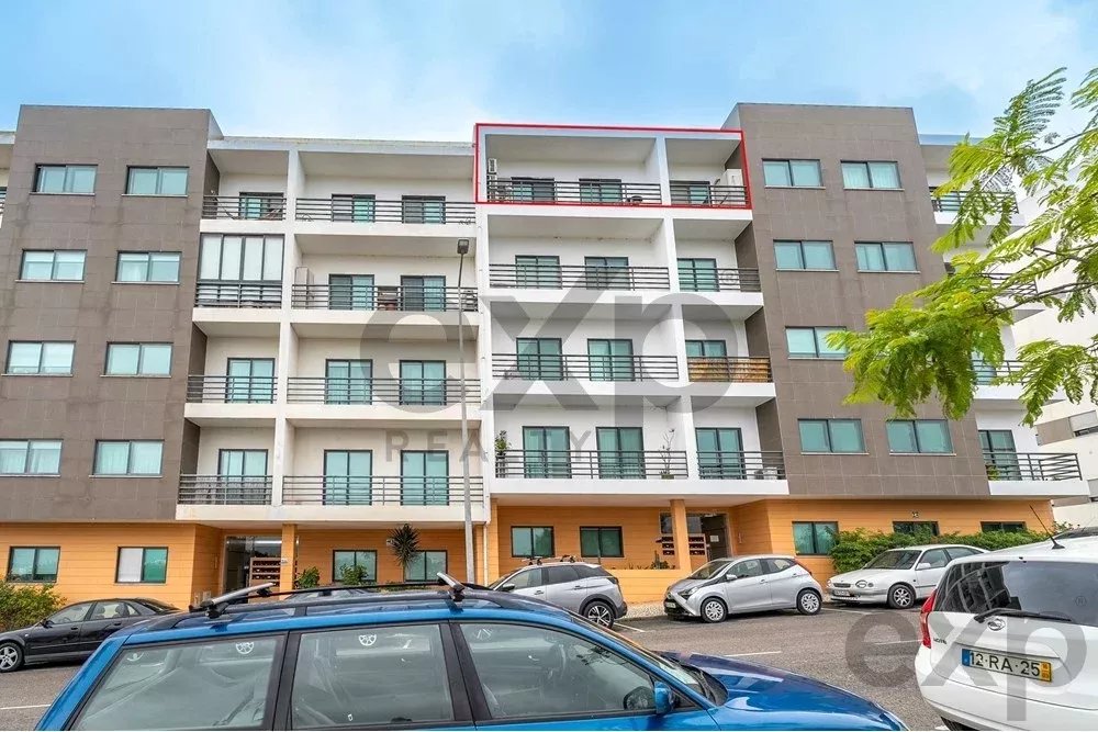 Multi-story apartment building with white balconies and gray facade, orange ground level, cars parked in front.