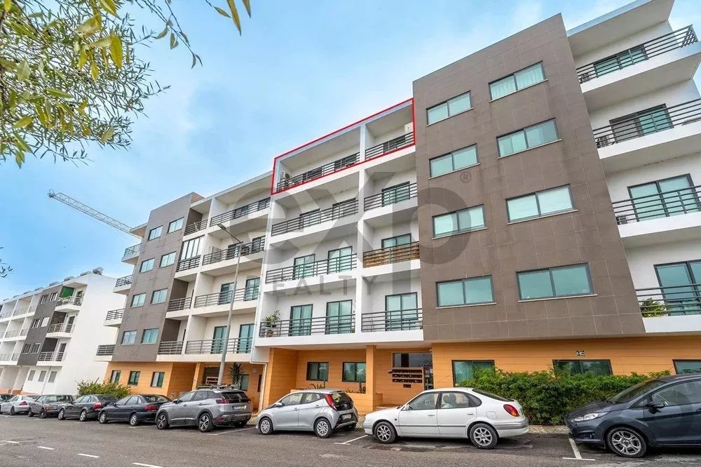 Modern multi-story apartment building with balconies, gray/brown facade, and a row of parked cars along the street under a blue sky.