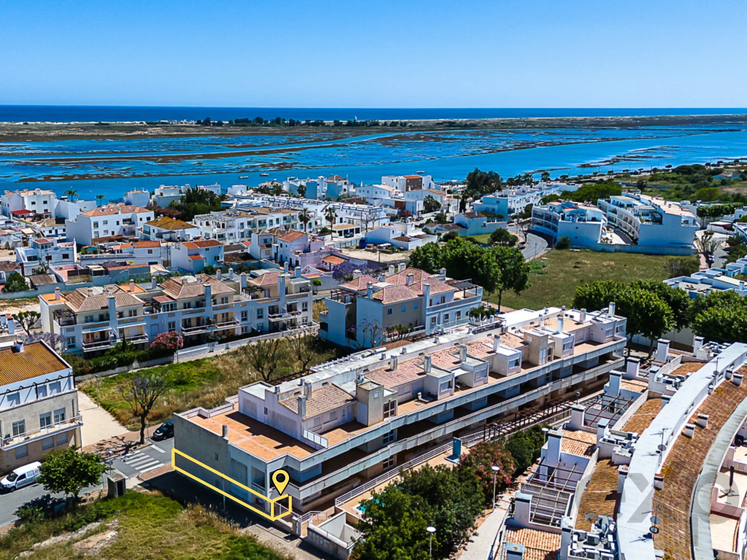 Aerial view of a coastal town with white houses along the waterfront and a yellow outlined building highlighted by a map pin marker.