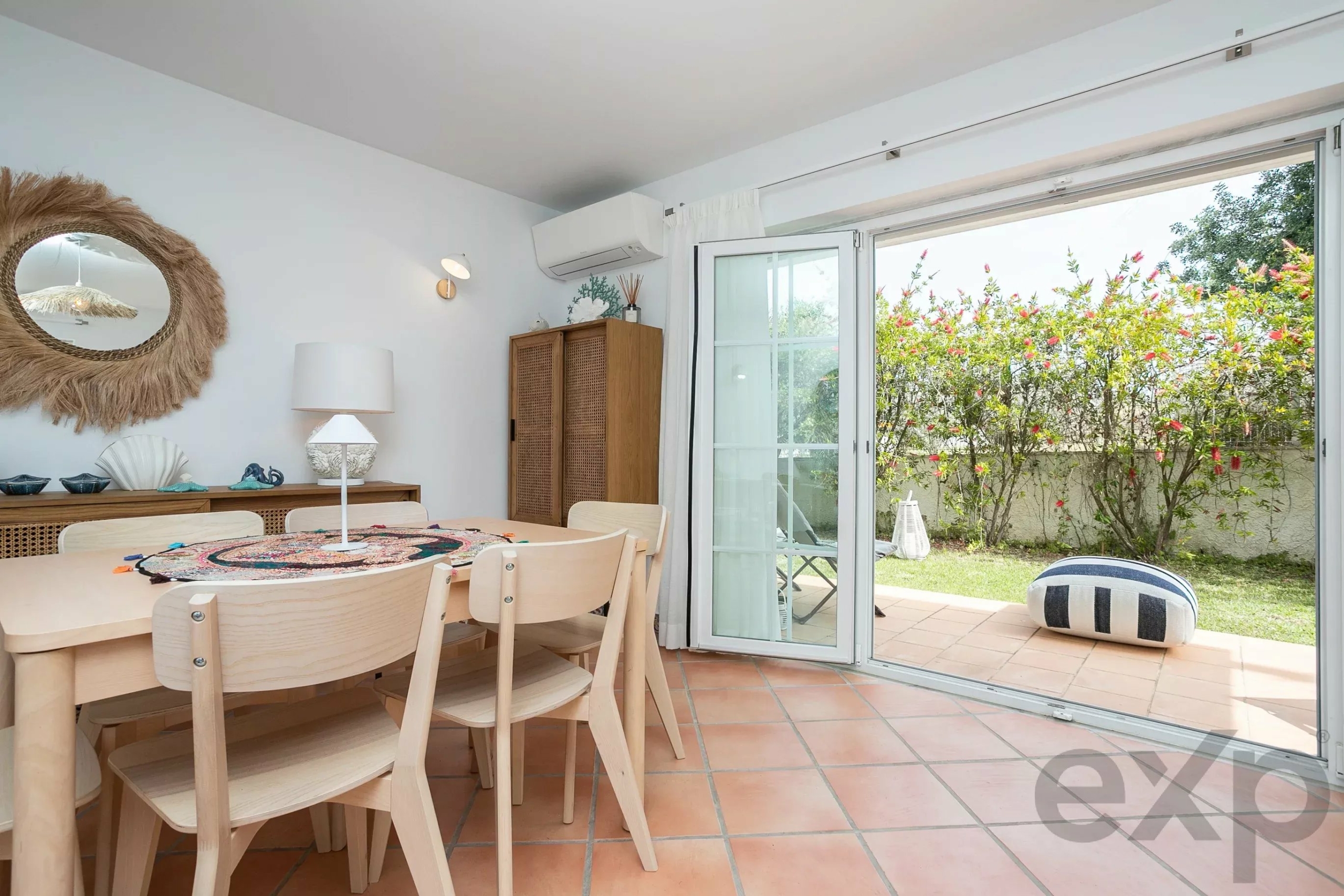 Bright dining area with wooden chairs around a round table, open glass doors to a garden with pink flowering bushes outside.