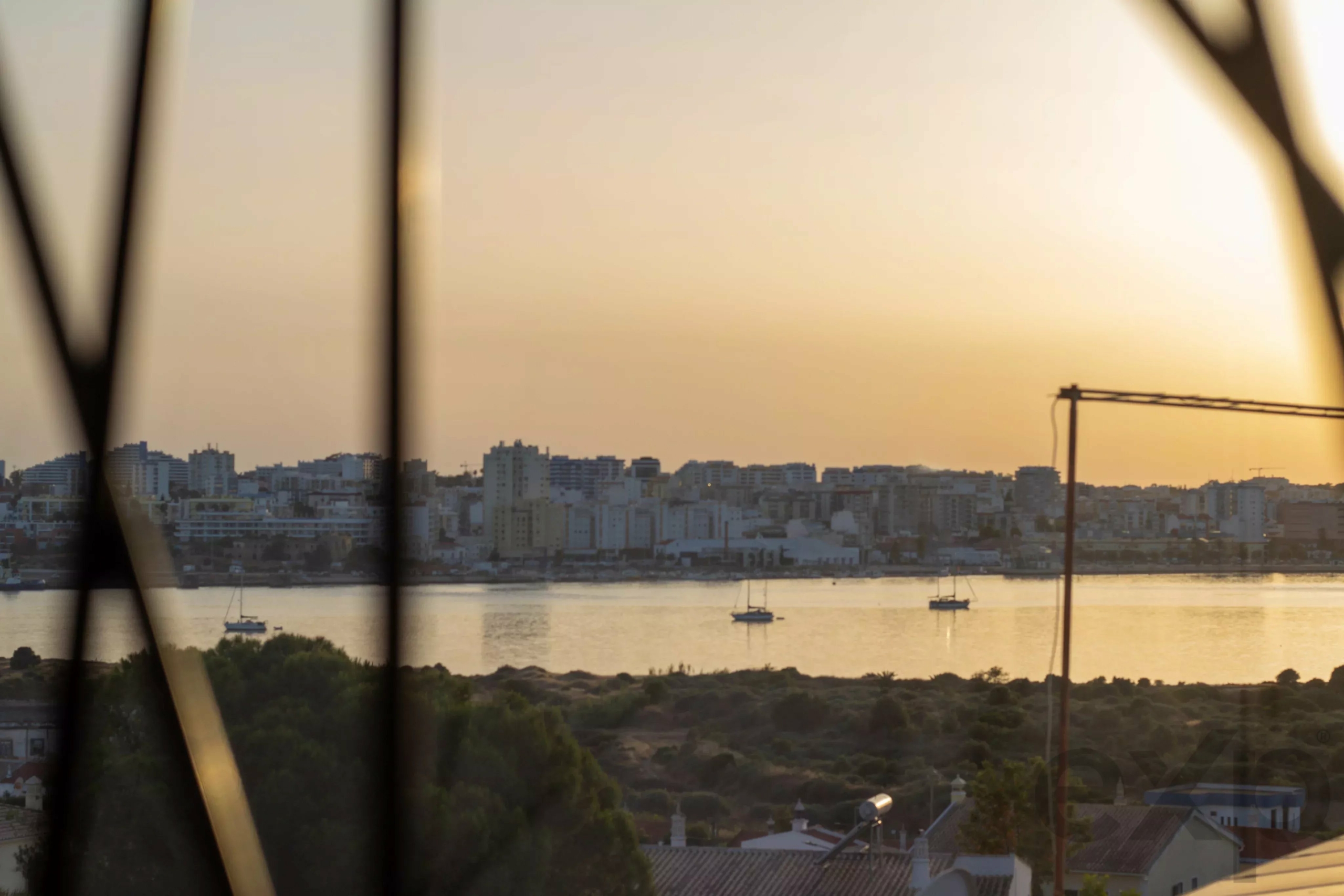 City skyline along a river at sunset, viewed through dark window blinds.