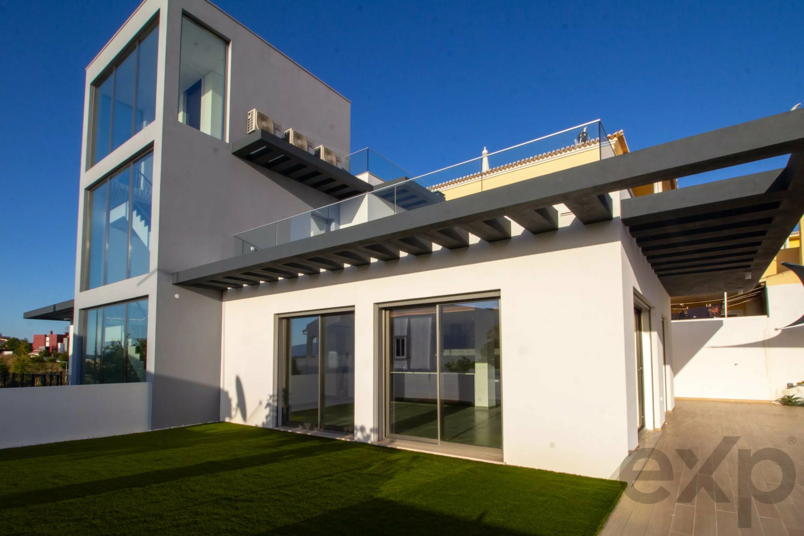 Modern two-story white house with large glass walls, glass railing terrace, and a neatly mowed lawn under a blue sky.