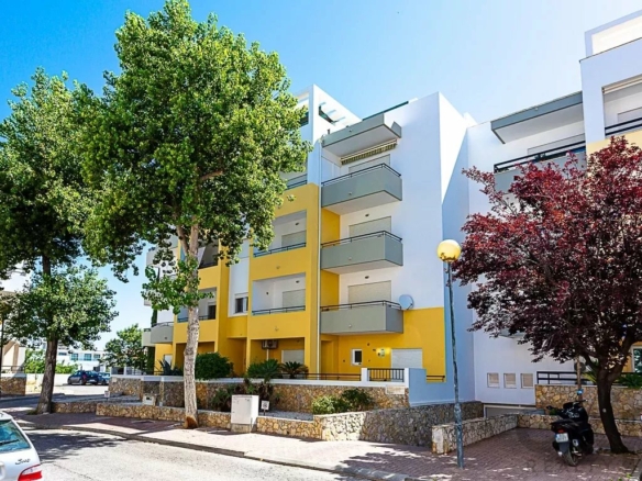 Modern white and yellow apartment building with multiple balconies on a tree-lined street; a scooter and cars parked nearby.