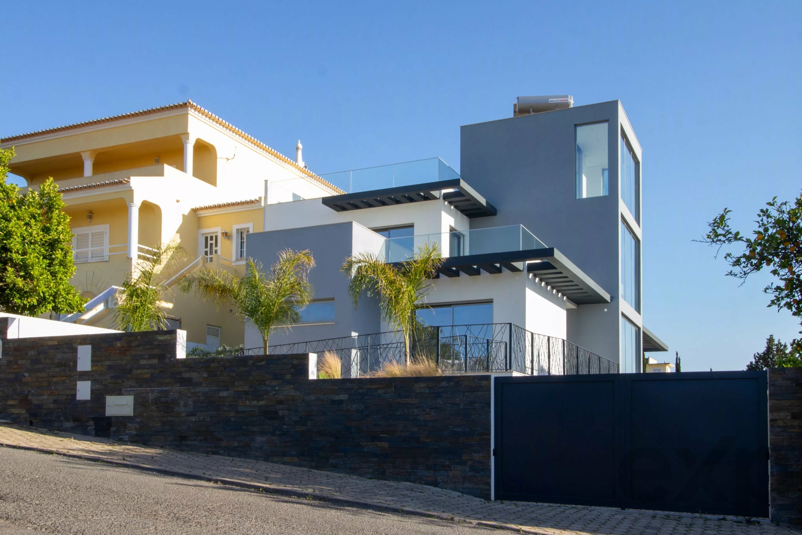 Modern residential building with yellow and gray façades, glass balconies, and palm trees against a clear blue sky, lower stone wall in foreground.