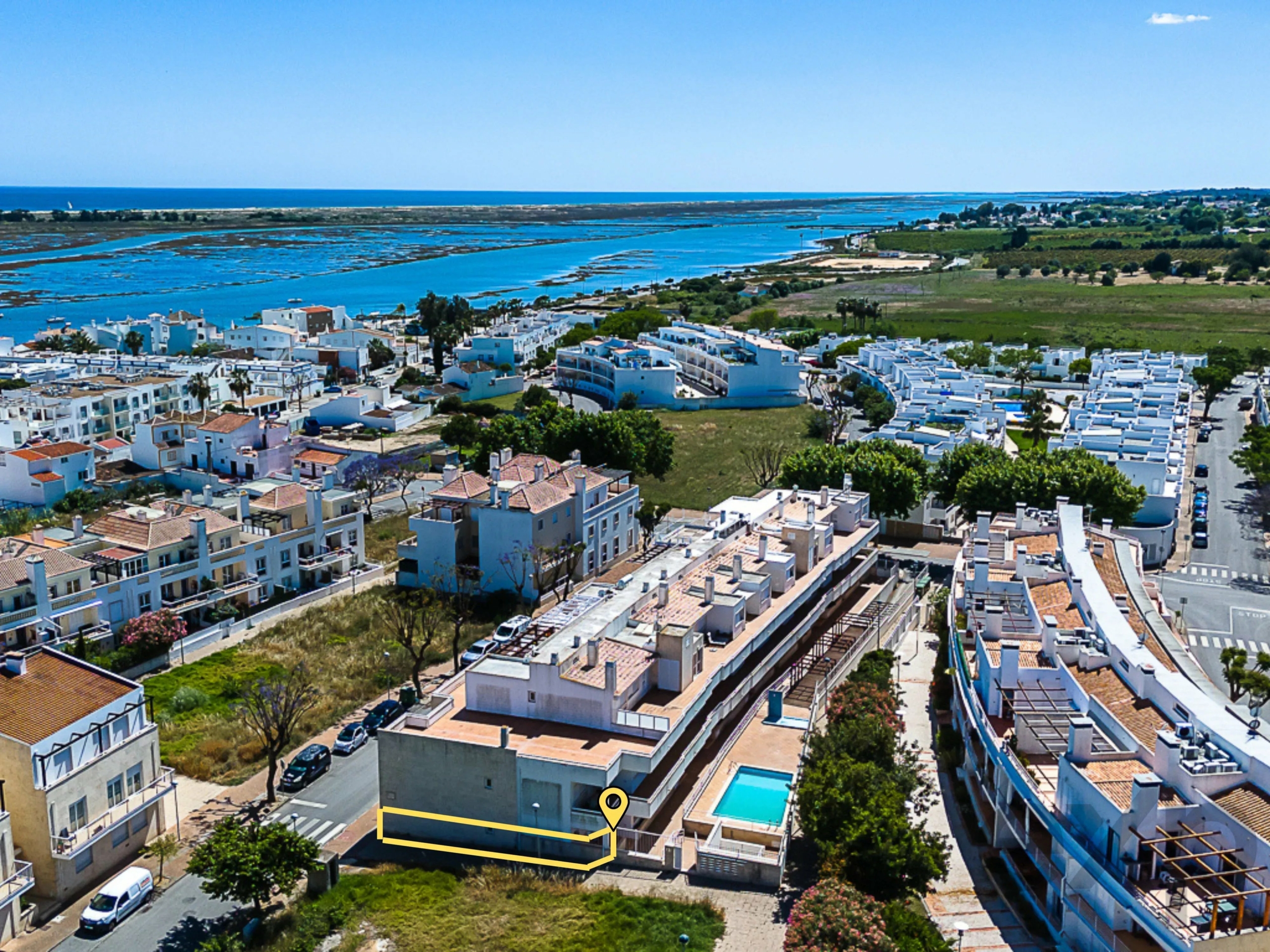 Aerial view of a seaside town with white buildings along streets and blue sea on the horizon; a yellow outlined property and location pin highlight a parcel in the foreground.