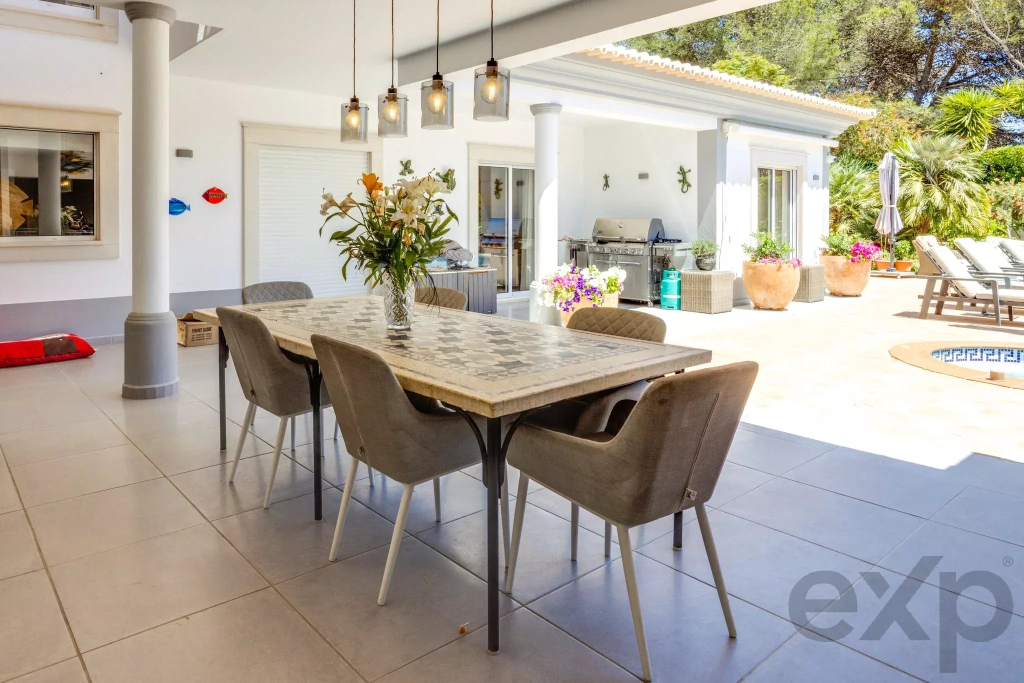 Outdoor patio with a rectangular dining table and brown chairs, white walls, and a built-in grill area under a shaded veranda. A vase of lilies on the table and potted plants line the space, with lounge chairs and a pool edge in the background.