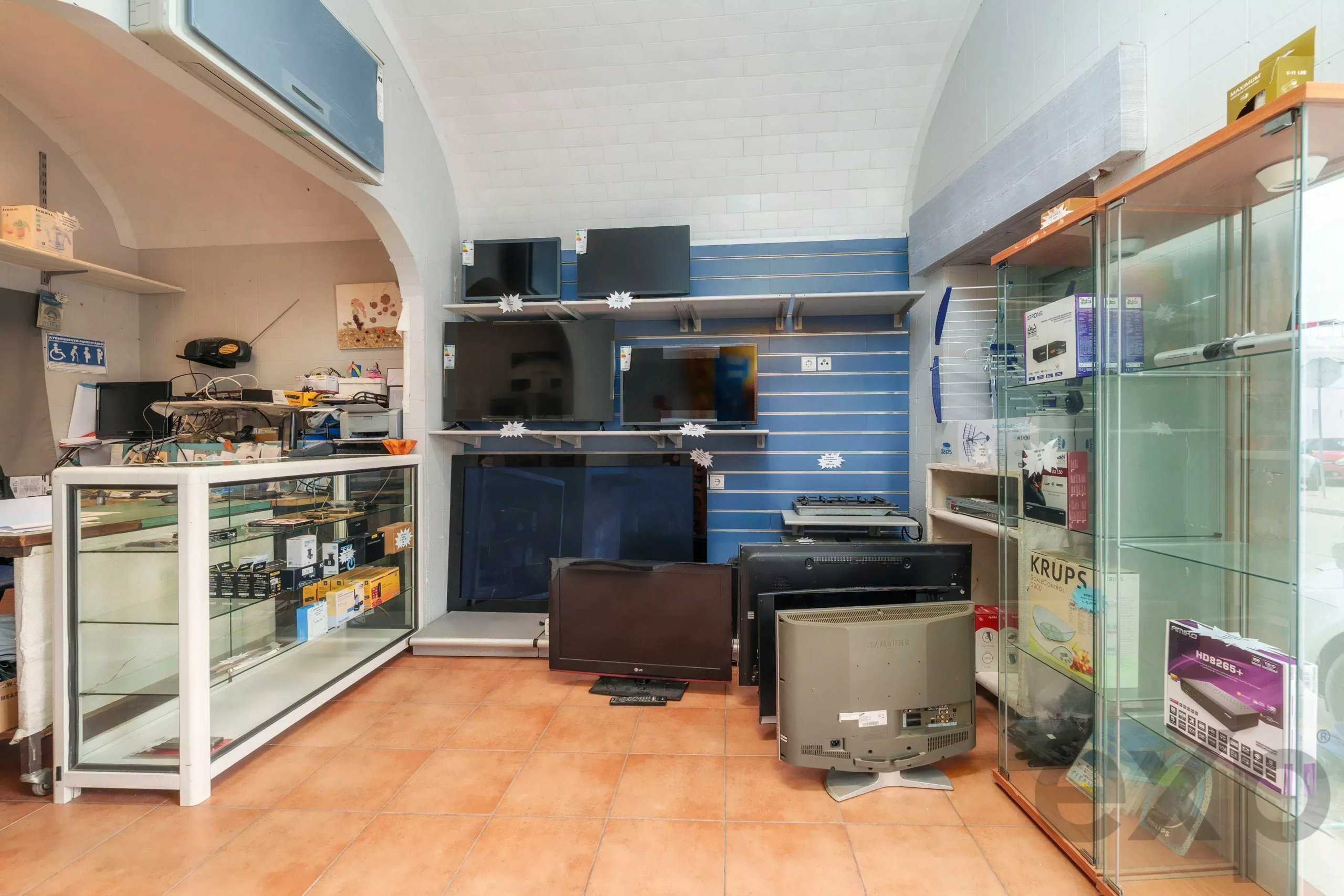 Electronics store interior with several wall-mounted TVs on blue slatwall shelves and larger TVs on the floor, plus glass display cases against the right wall.