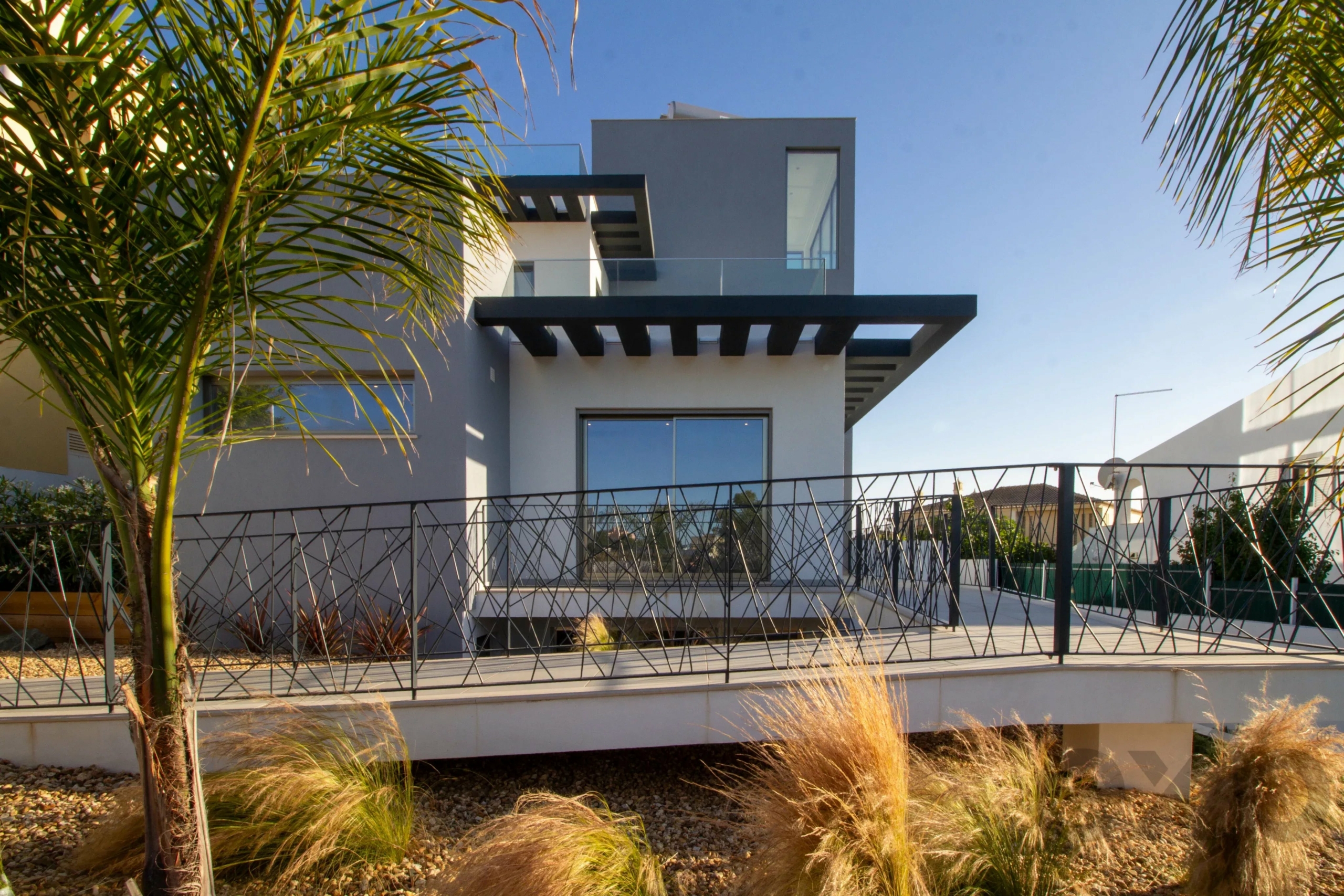 Modern white multi-story house with glass balconies framed by palm trees against a blue sky, surrounded by a metal fence and desert landscaping.