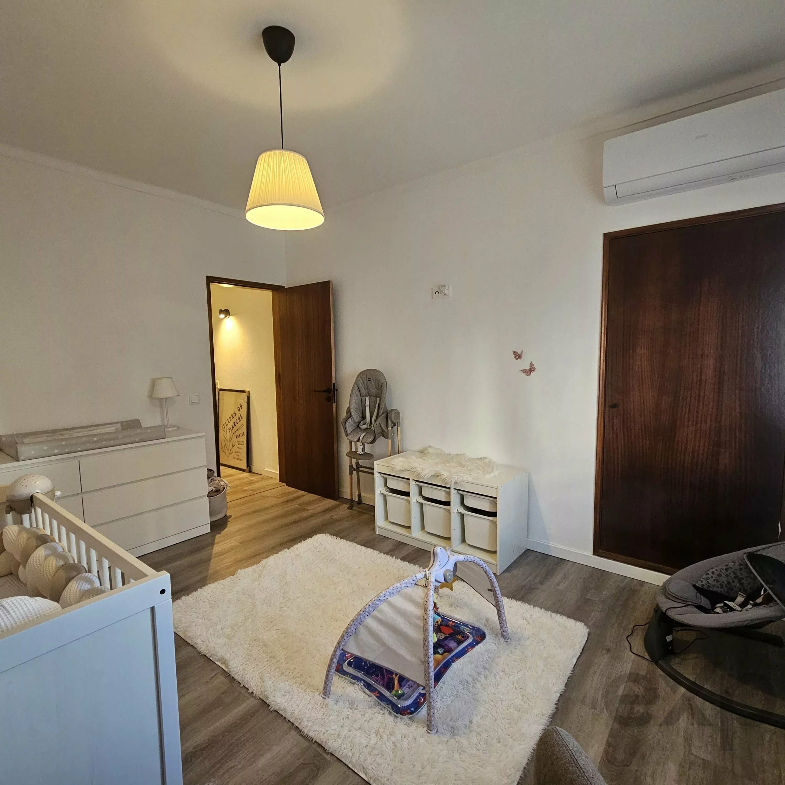 cozy nursery with white furniture (crib and chest), cream shag rug, rocking chair, butterfly wall decals, and a warm ceiling pendant light; door open to adjacent room