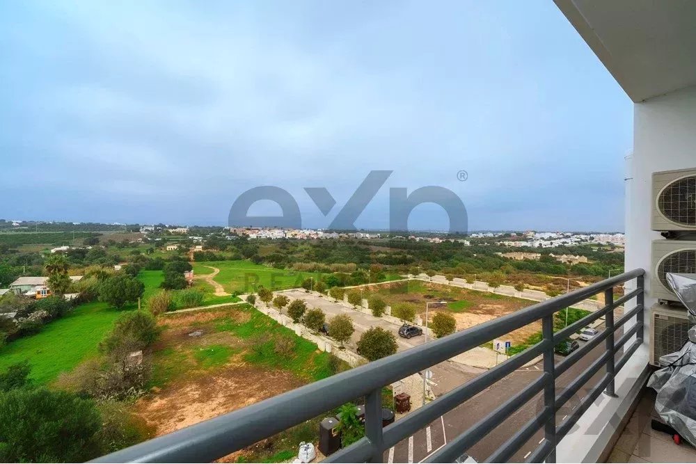 Balcony view of green fields and a distant town under a cloudy sky, with a metal railing in the foreground.