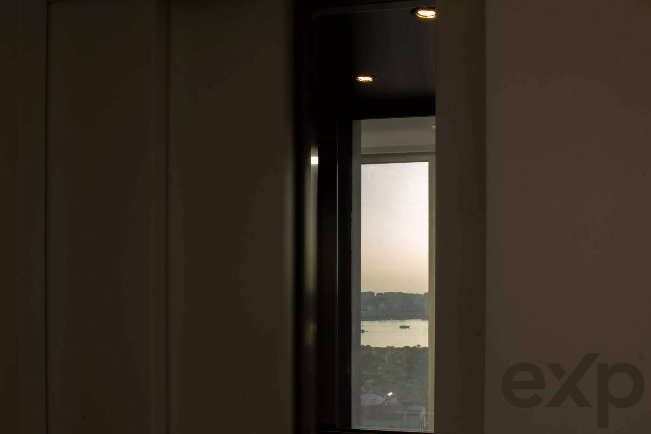 Dim interior with a narrow glass window showing a distant, sunlit landscape outside at dusk.